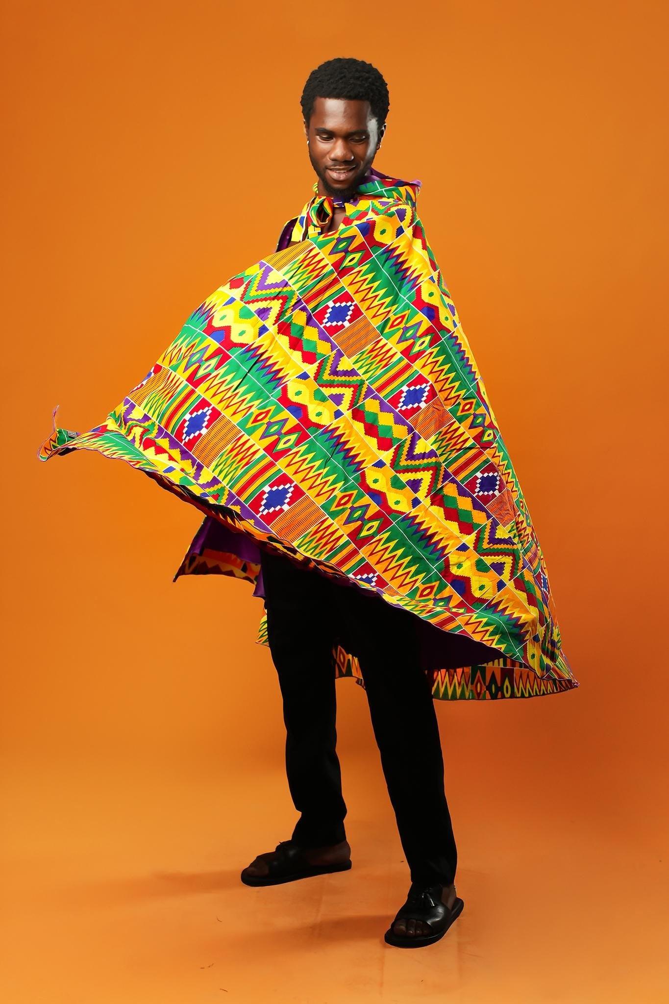 Man standing against an orange background wearing a purple festival cape with a colourful Kente African print trim. The cape ties at the neck with matching Kente fabric.