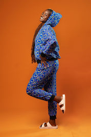 Woman standing against an orange background wearing blue African print jogger trousers with a black drawstring waistband and black ankle cuffs. The trousers feature a colourful geometric pattern in blue, yellow, and red.