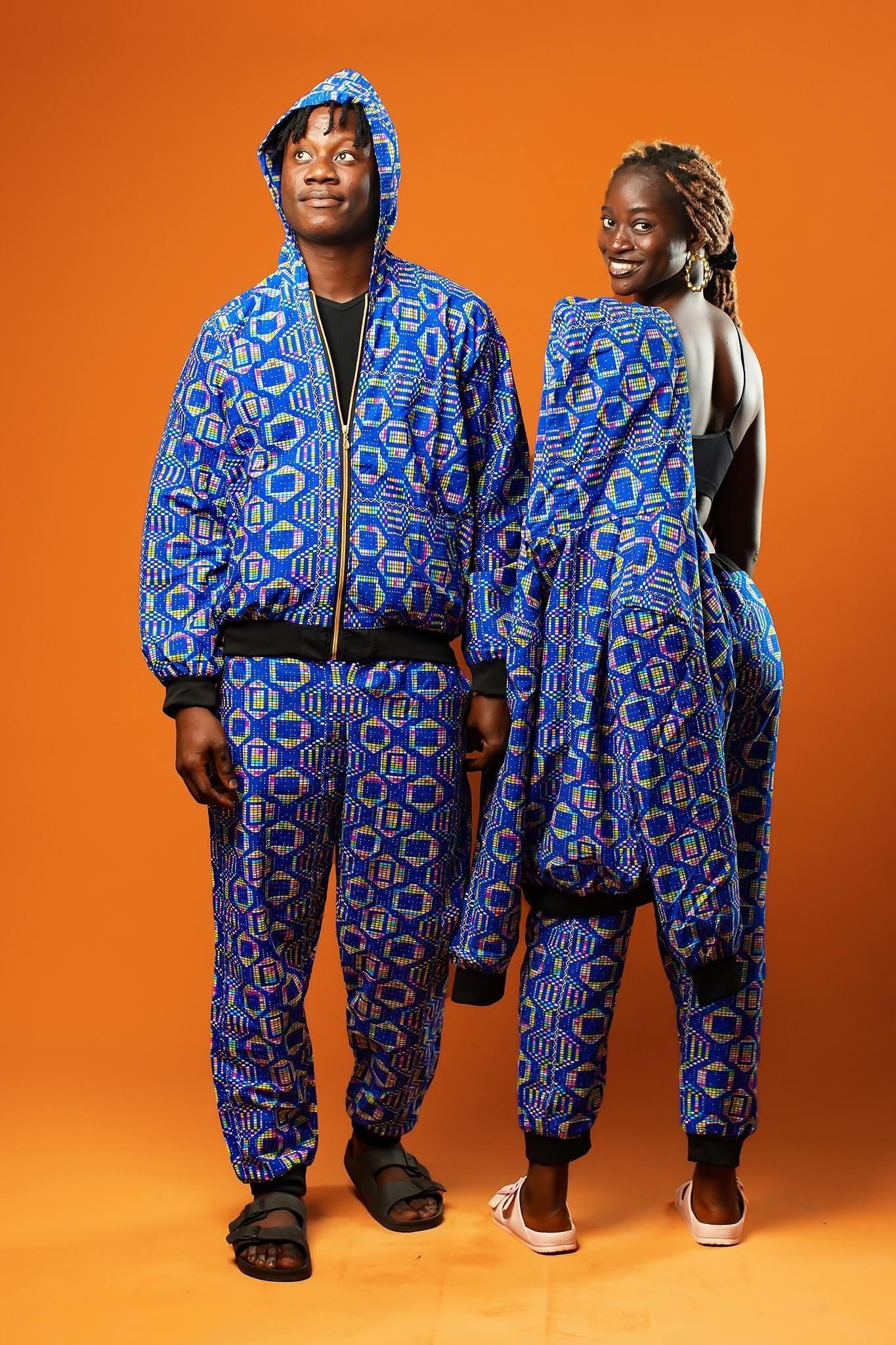 Man and woman standing against an orange background wearing matching African print tracksuits. The outfits are made from blue cotton fabric with a colourful geometric pattern in yellow, red, and green. The man wears a full zip-up hoodie and jogger trousers with black cuffs and waistband. The woman wears matching joggers and carries the hoodie over her shoulder.