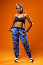 Woman standing against an orange background wearing blue African print jogger trousers with a black drawstring waistband and black ankle cuffs. The trousers feature a colourful geometric pattern in blue, yellow, and red.