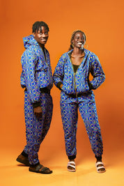 Man and woman standing against an orange background wearing matching African print tracksuits. The outfits are made from blue cotton fabric with a colourful geometric pattern in yellow, red, and green. The man wears a full zip-up hoodie and jogger trousers with black cuffs and waistband. The woman wears matching joggers and hoodie