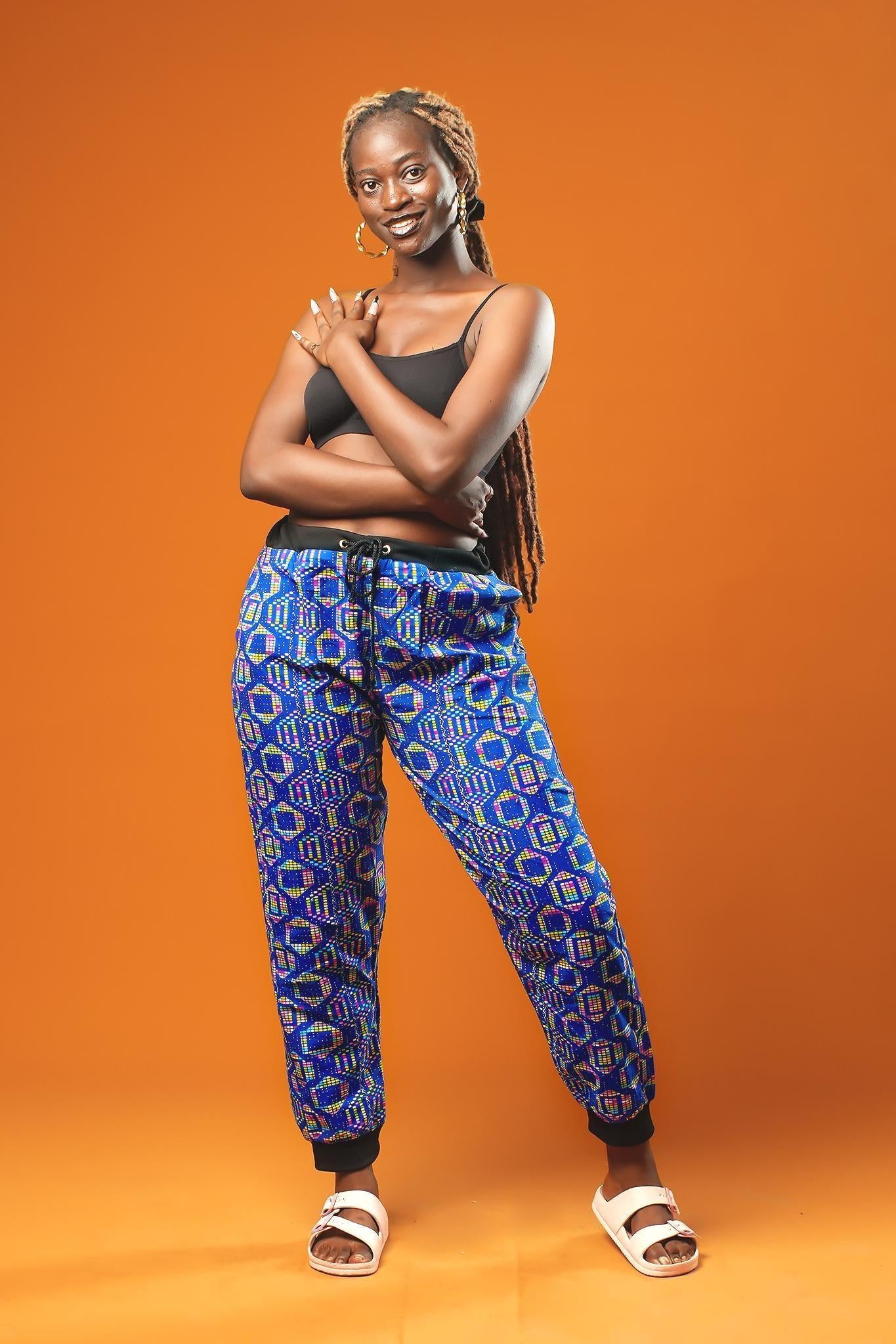 Woman standing against an orange background wearing blue African print jogger trousers with a black drawstring waistband and black ankle cuffs. The trousers feature a colourful geometric pattern in blue, yellow, and red.
