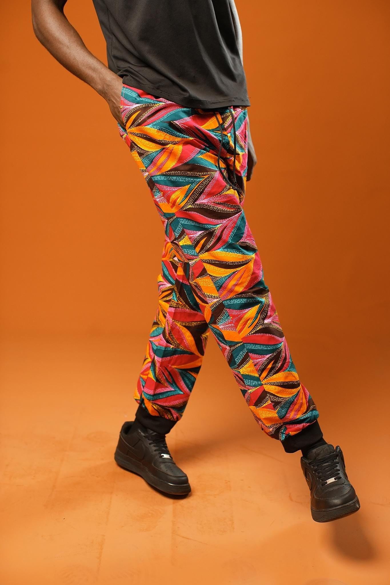 Man standing against an orange background wearing colourful African Ankara trousers made from bright orange, pink, green, and yellow tribal print fabric. The trousers have a relaxed fit with a black drawstring waistband and black ankle cuffs.