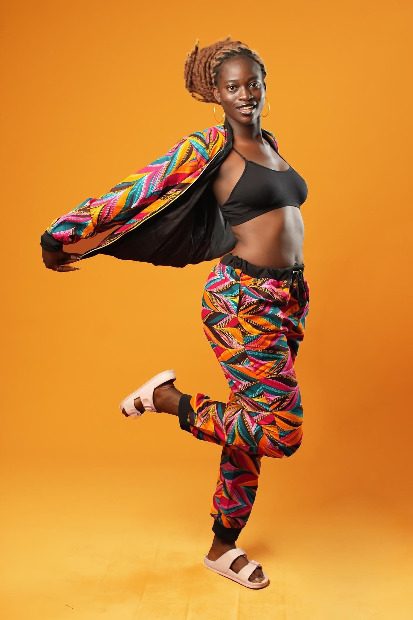 Woman standing against an orange background wearing colourful African Ankara trousers made from bright orange, pink, green, and yellow tribal print fabric. The trousers have a relaxed fit with a black drawstring waistband and black ankle cuffs. Styled as part of a matching tracksuit set.