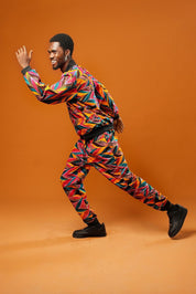 Man standing against an orange background wearing colourful African Ankara trousers made from bright orange, pink, green, and yellow tribal print fabric. The trousers have a relaxed fit with a black drawstring waistband and black ankle cuffs. Styled as part of a matching tracksuit set.