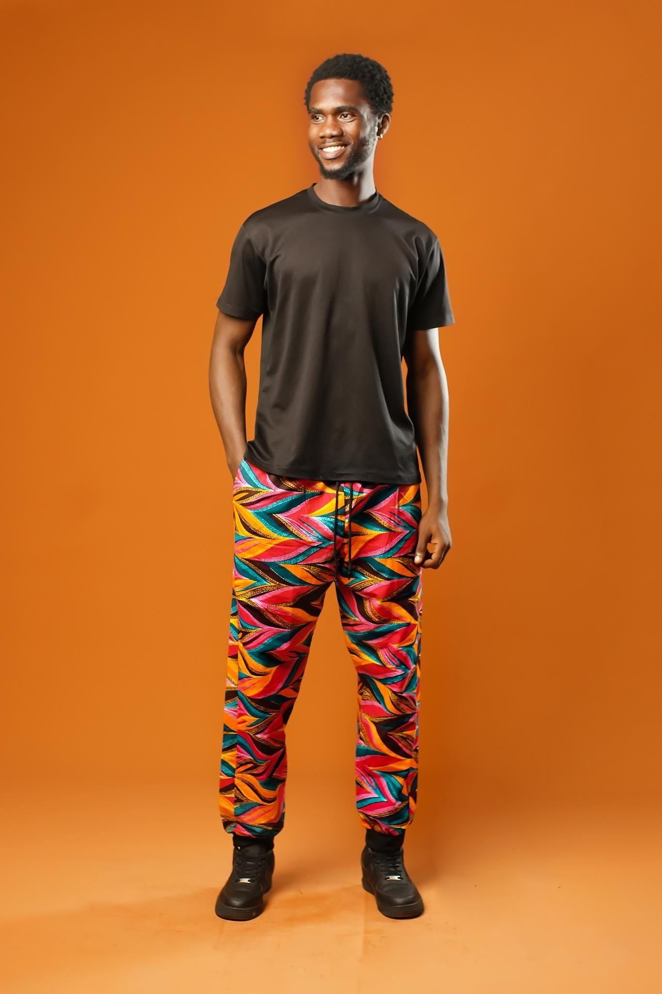 Man standing against an orange background wearing colourful African Ankara trousers made from bright orange, pink, green, and yellow tribal print fabric. The trousers have a relaxed fit with a black drawstring waistband and black ankle cuffs.