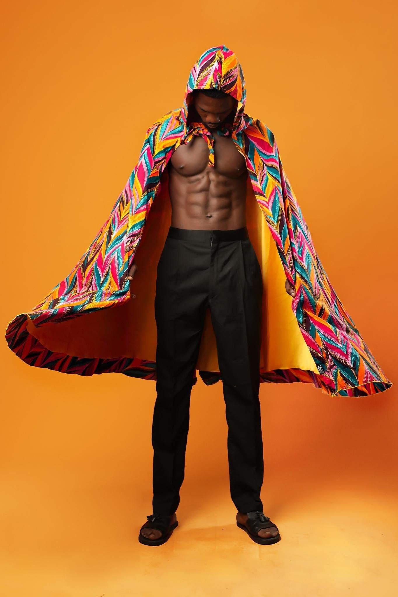 Man standing against an orange background wearing a colourful African print festival cape with a hood. The outside of the unisex cape has a multicoloured feather-like pattern, and the inside is lined with shiny yellow fabric. The cape ties at the neck with matching fabric.