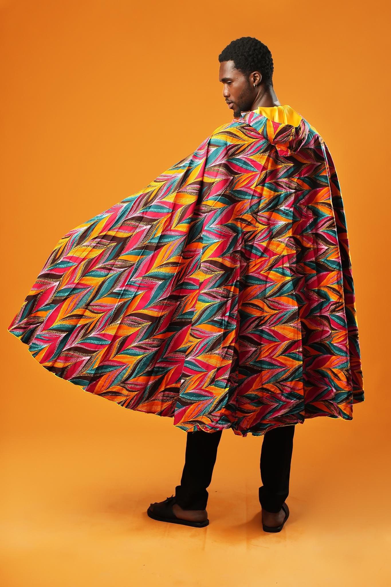 Man standing against an orange background wearing a colourful African print festival cape with a hood. The outside of the unisex cape has a multicoloured feather-like pattern, and the inside is lined with shiny yellow fabric. The cape ties at the neck with matching fabric.