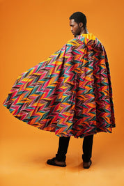 Man standing against an orange background wearing a colourful African print festival cape with a hood. The outside of the unisex cape has a multicoloured feather-like pattern, and the inside is lined with shiny yellow fabric. The cape ties at the neck with matching fabric.