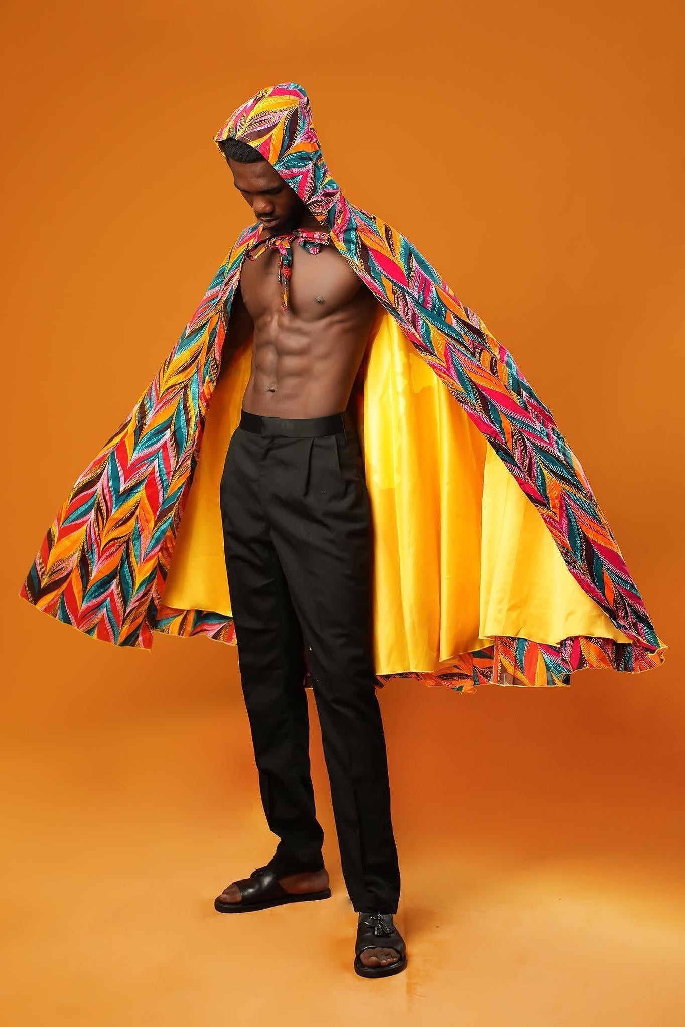 Man standing against an orange background wearing a colourful African print festival cape with a hood. The outside of the unisex cape has a multicoloured feather-like pattern, and the inside is lined with shiny yellow fabric. The cape ties at the neck with matching fabric.