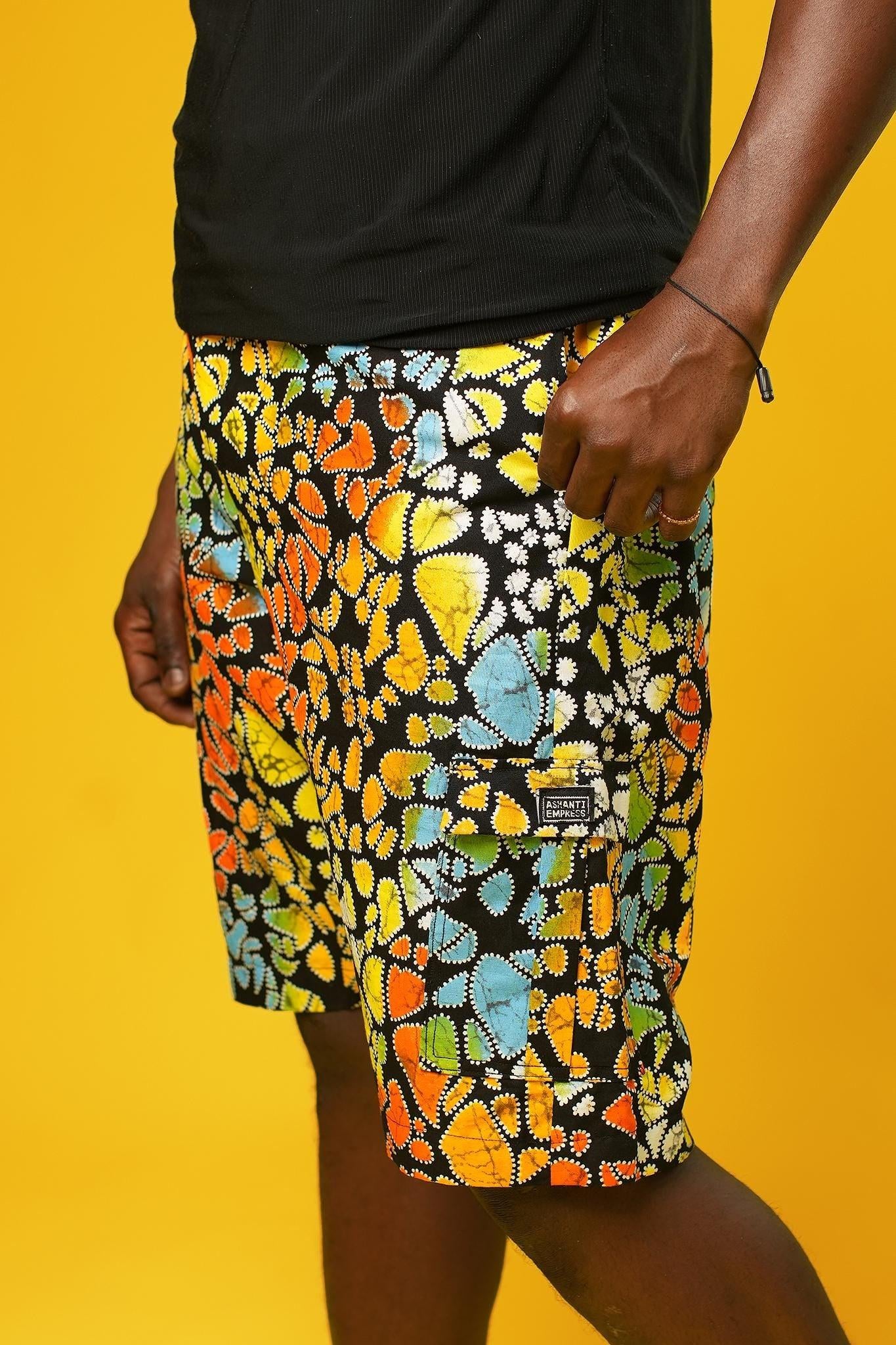 A man stands in front of a yellow background wearing Funky Men’s Cargo Shorts made from multicoloured African print fabric with a butterfly pattern in orange, yellow, blue, and black. The shorts have a relaxed fit and side pockets, paired with a matching short-sleeve shirt. This is a bold Summer Hawaiian Shorts look, ideal for African Print Men’s Clothing, a Hippie Festival Set, or a Carnival Outfit from Ashanti Empress.