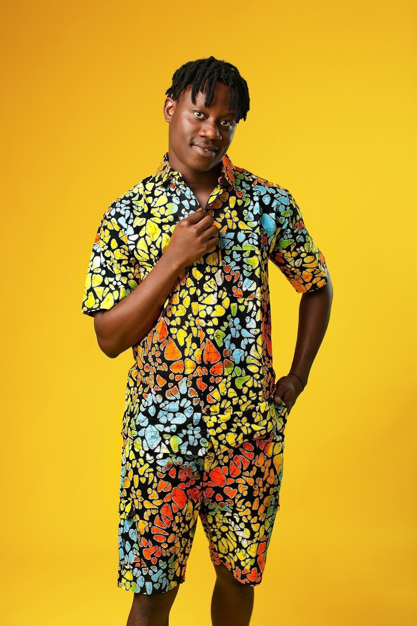 A man stands confidently against a bright yellow background wearing a vibrant matching two-piece outfit made from multicoloured African wax print fabric. The set features a short-sleeved button-up shirt and shorts, both covered in a bold, abstract pattern of orange, yellow, blue, green, and red shapes outlined in black.