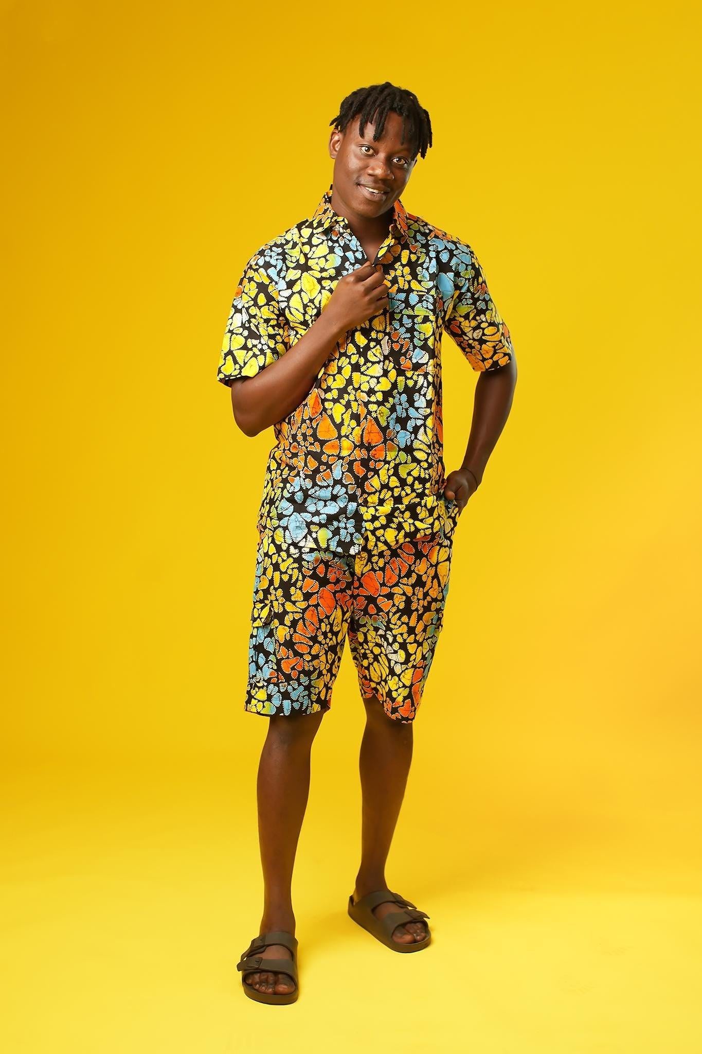 A man stands confidently against a bright yellow background wearing a vibrant matching two-piece outfit made from multicoloured African wax print fabric. The set features a short-sleeved button-up shirt and shorts, both covered in a bold, abstract pattern of orange, yellow, blue, green, and red shapes outlined in black.