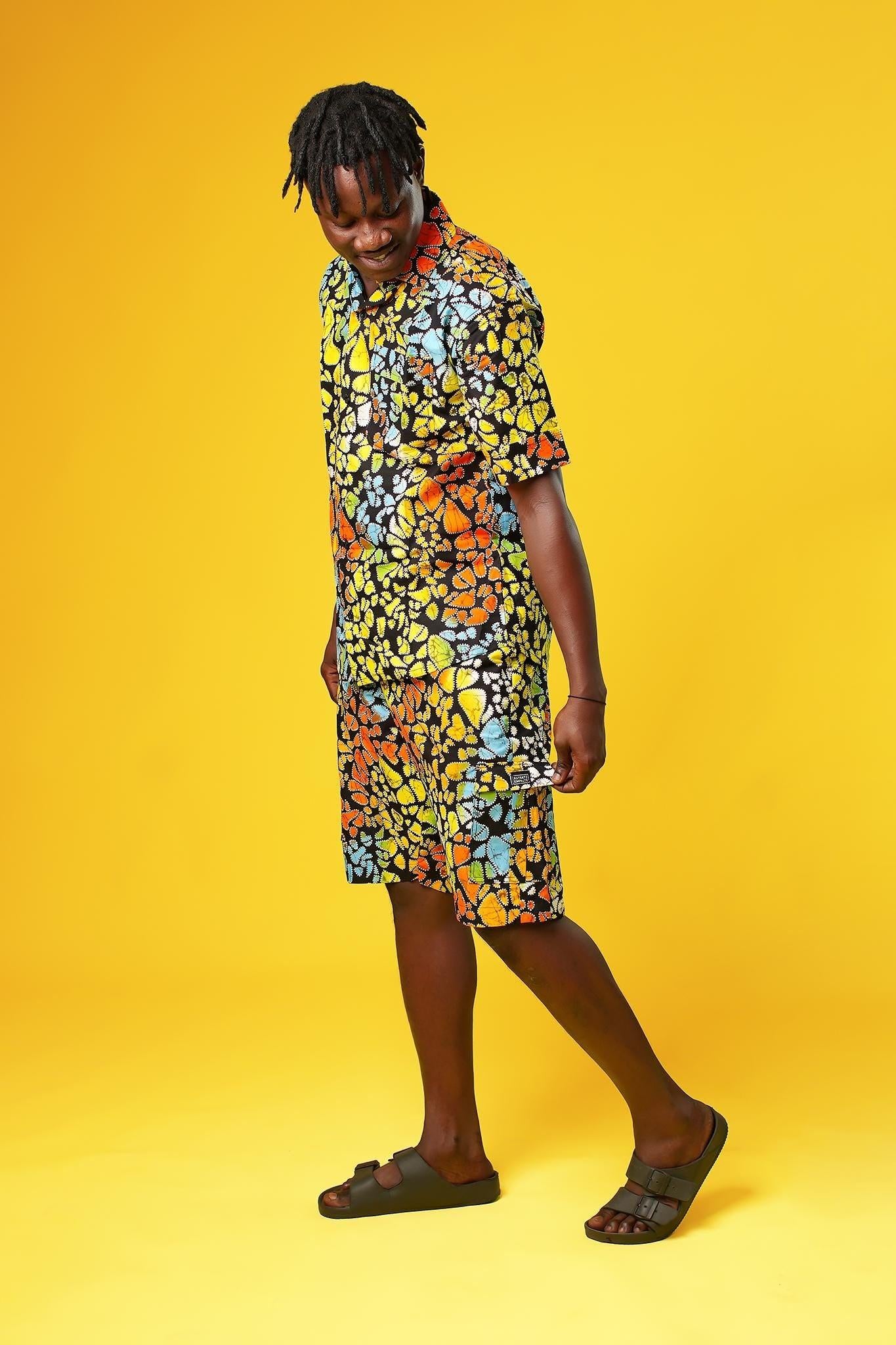 A man stands confidently against a bright yellow background wearing a vibrant matching two-piece outfit made from multicoloured African wax print fabric. The set features a short-sleeved button-up shirt and shorts, both covered in a bold, abstract pattern of orange, yellow, blue, green, and red shapes outlined in black.
