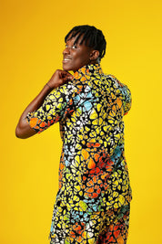A man stands confidently against a bright yellow background wearing a vibrant matching two-piece outfit made from multicoloured African wax print fabric. The set features a short-sleeved button-up shirt and shorts, both covered in a bold, abstract pattern of orange, yellow, blue, green, and red shapes outlined in black.