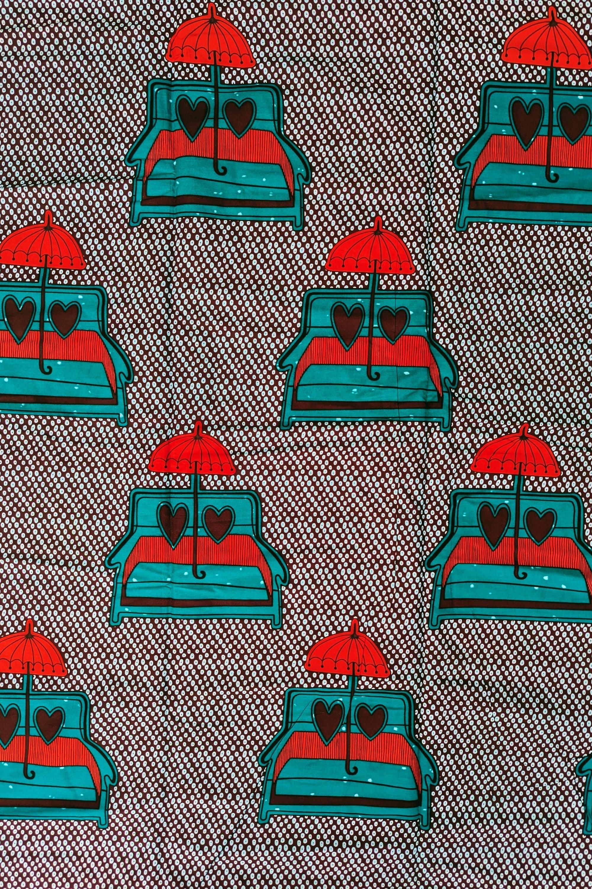 African wax print fabric featuring a playful and romantic design of turquoise blue sofas with red and brown heart-shaped pillows, each topped with a bright red umbrella. The background is a dark brown covered in a pattern of small white teardrop shapes, creating a bold and eye-catching contrast. Perfect for statement fashion or unique home decor projects.