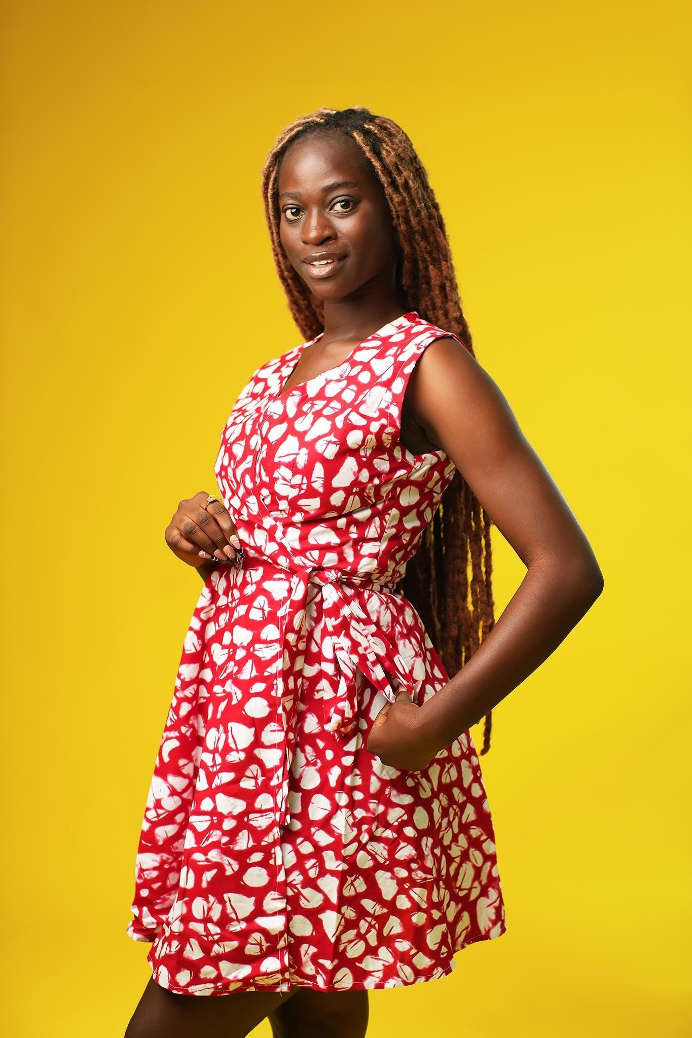 Woman wearing a red and white abstract batik-style African print wrap dress. The dress is sleeveless with a V-neckline and tie waist, made from cotton fabric, and falls above the knees.
