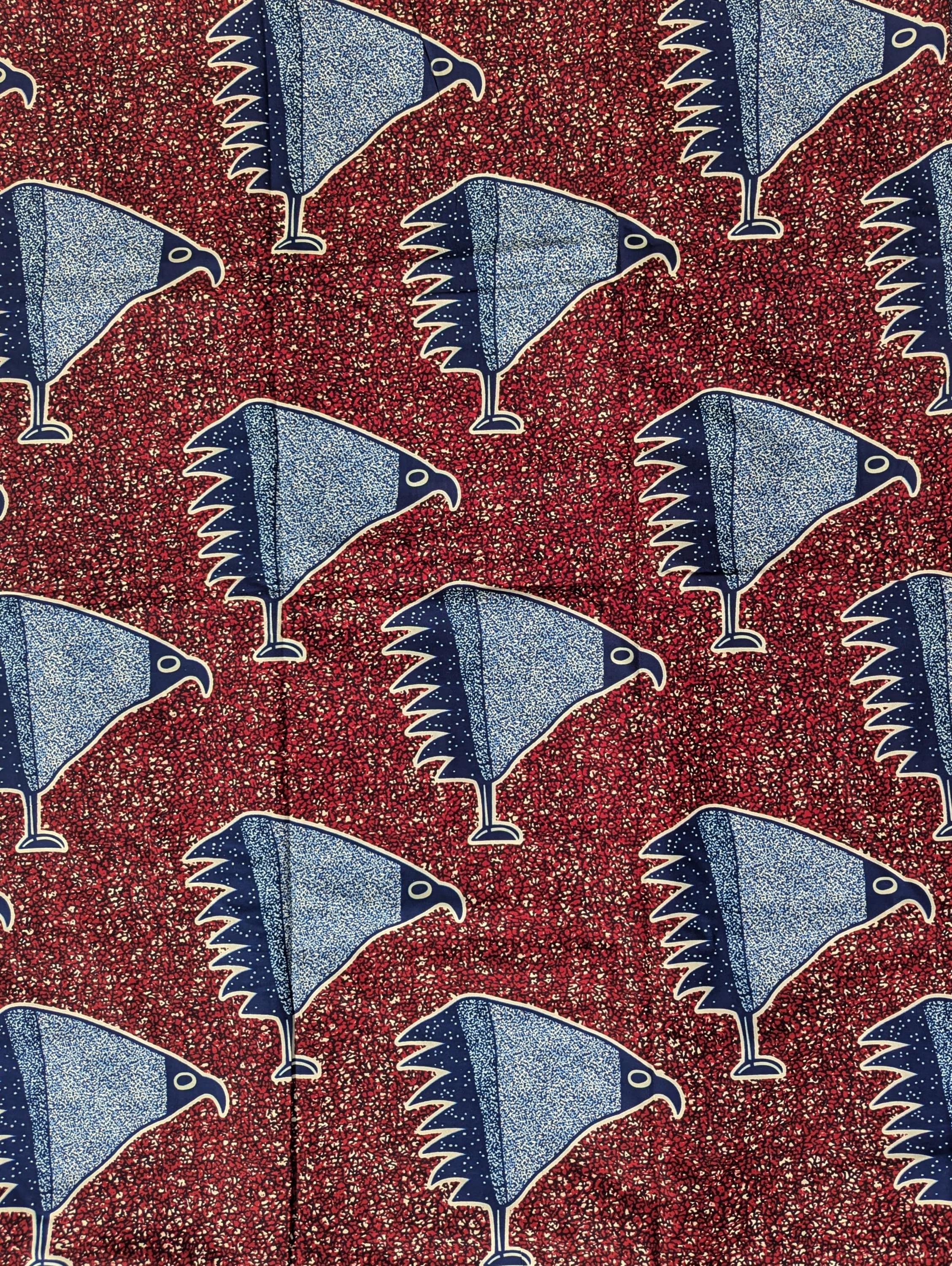 African wax print fabric with a striking pattern of stylised blue birds resembling eagles or hawks, each standing on one leg. The birds are illustrated in a speckled texture with navy and sky blue wings, set against a rich red background filled with small, abstract cream and black shapes. Bold and graphic, this fabric is ideal for statement fashion or unique accessories.