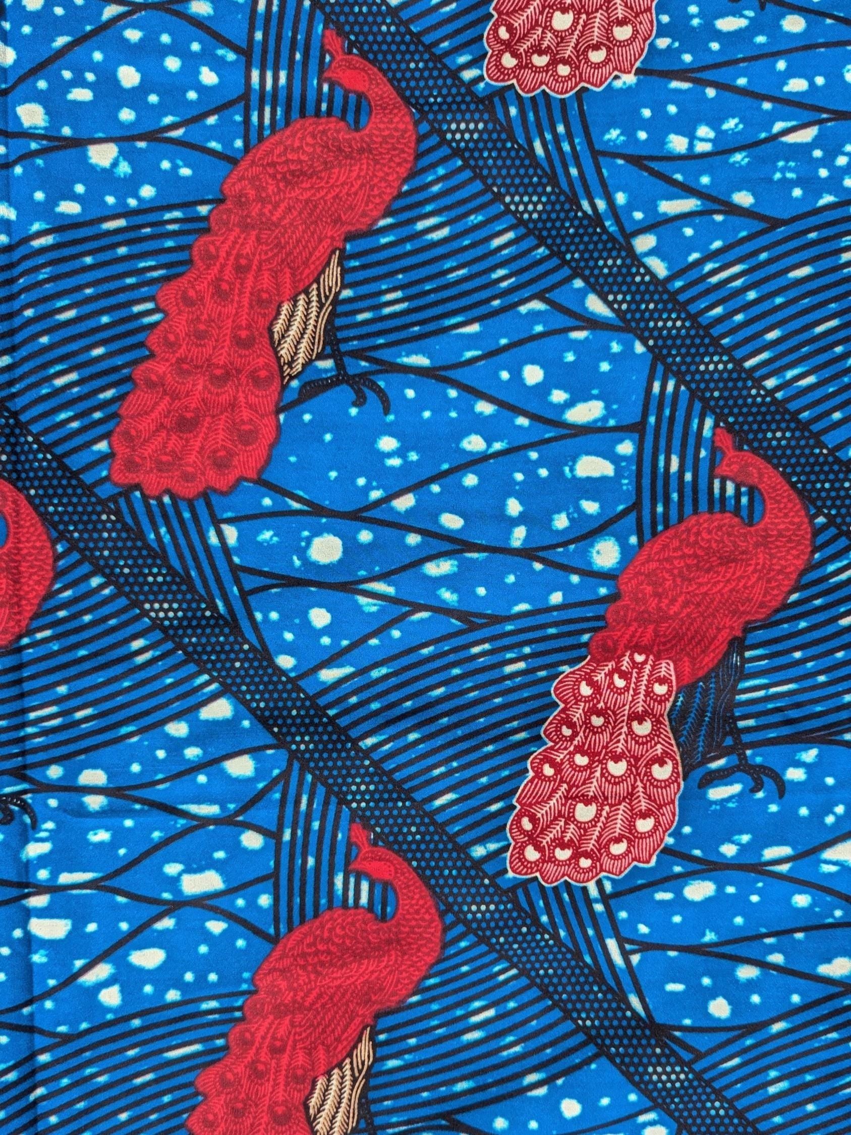 Bright and bold African wax print fabric featuring elegant pink peacocks with intricately patterned tail feathers standing against a vivid blue background. The design includes diagonal black lines and abstract wave-like patterns with splashes of cream, creating a striking contrast and sense of movement. Perfect for standout fashion pieces or statement accessories.