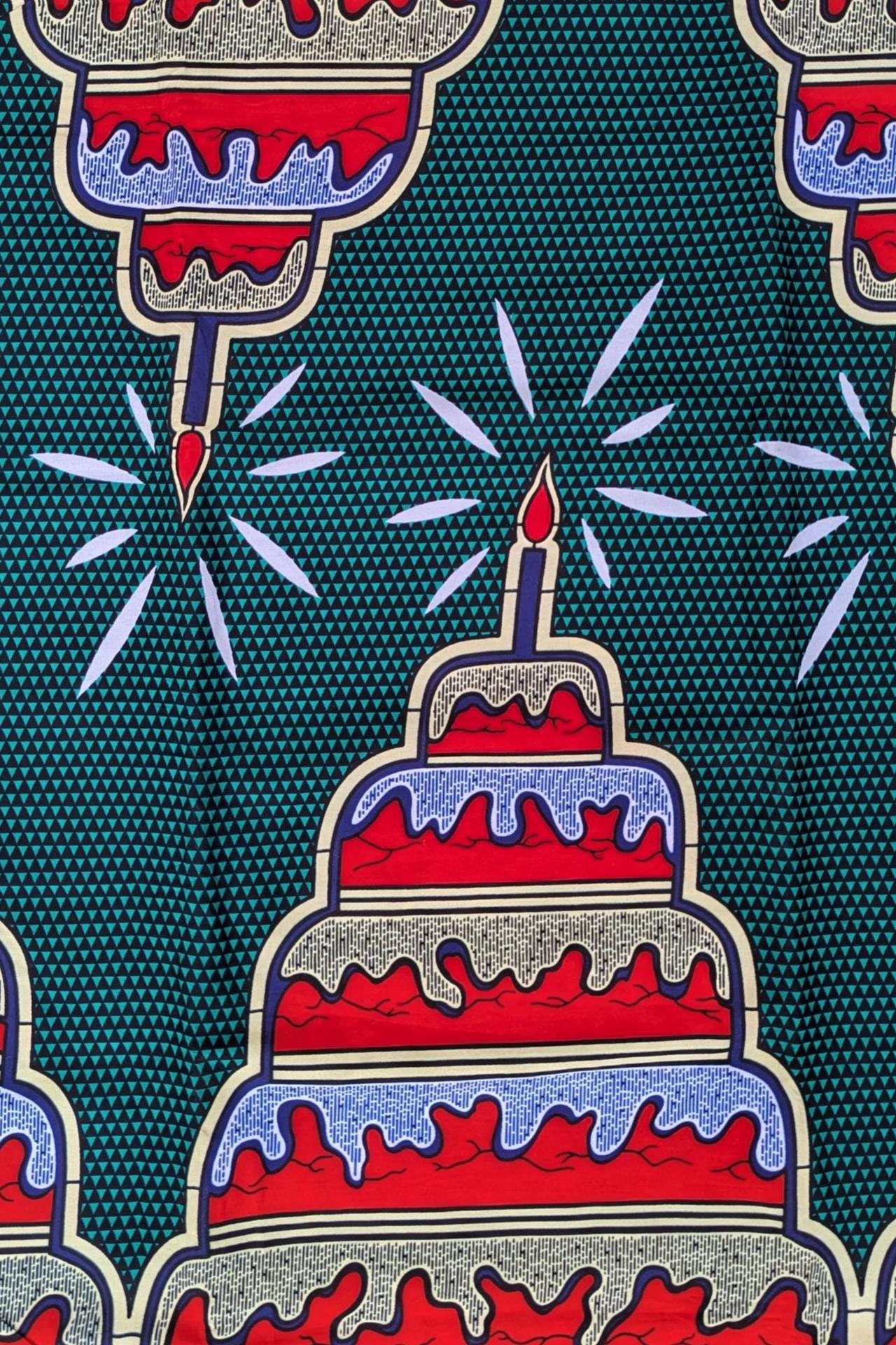 Bold and colourful African wax print fabric featuring a repeating pattern of tiered birthday cakes with red, white, and blue icing and a single lit candle on top. The background is a deep green with a small geometric triangle pattern, and the cakes are arranged upright and upside down in an alternating design.
