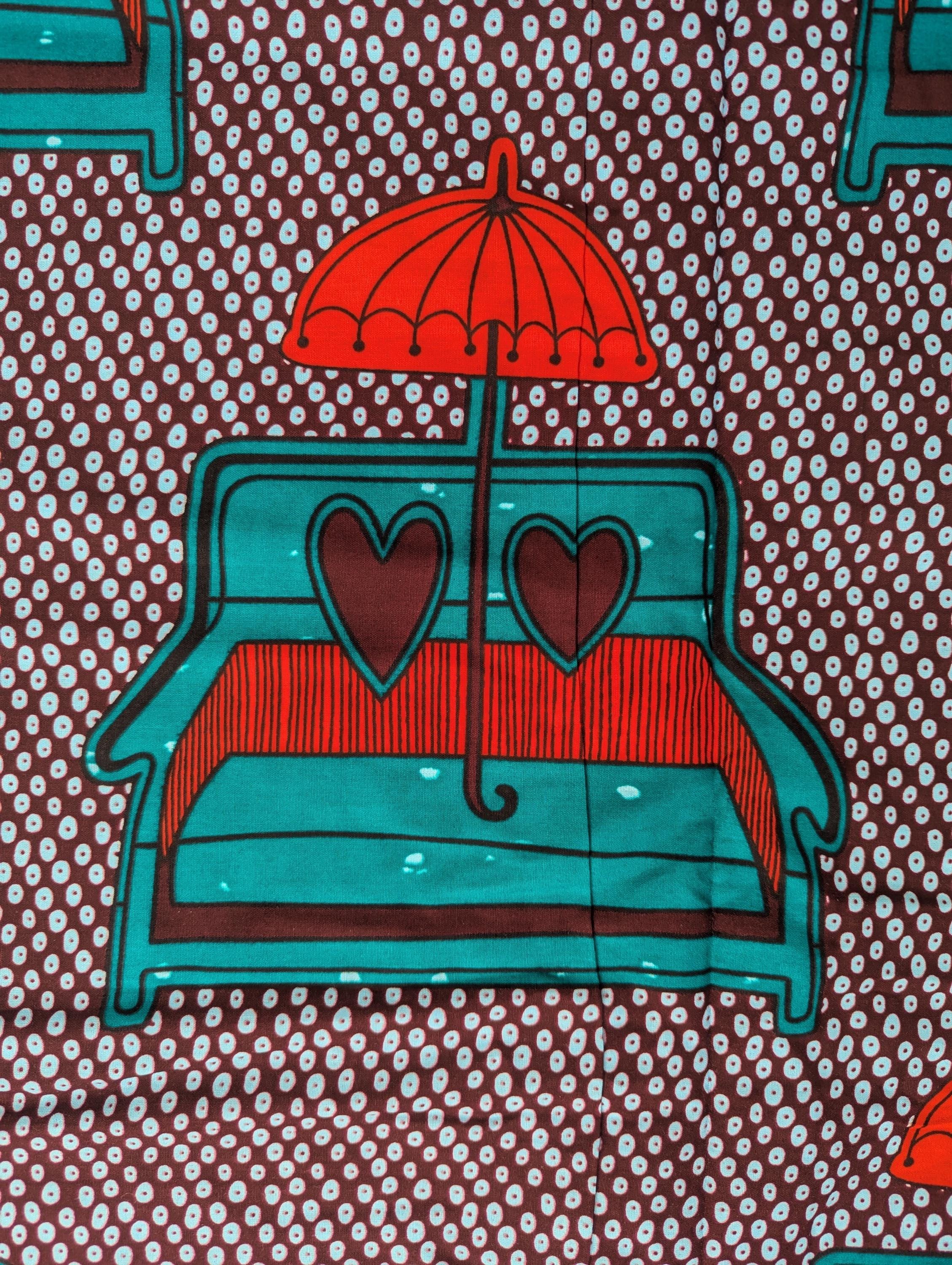 African wax print fabric featuring a playful and romantic design of turquoise blue love seats with red and brown heart-shaped pillows, each topped with a bright red umbrella. The background is a dark brown covered in a pattern of small white teardrop shapes, creating a bold and eye-catching contrast. Perfect for statement fashion or unique home decor projects.