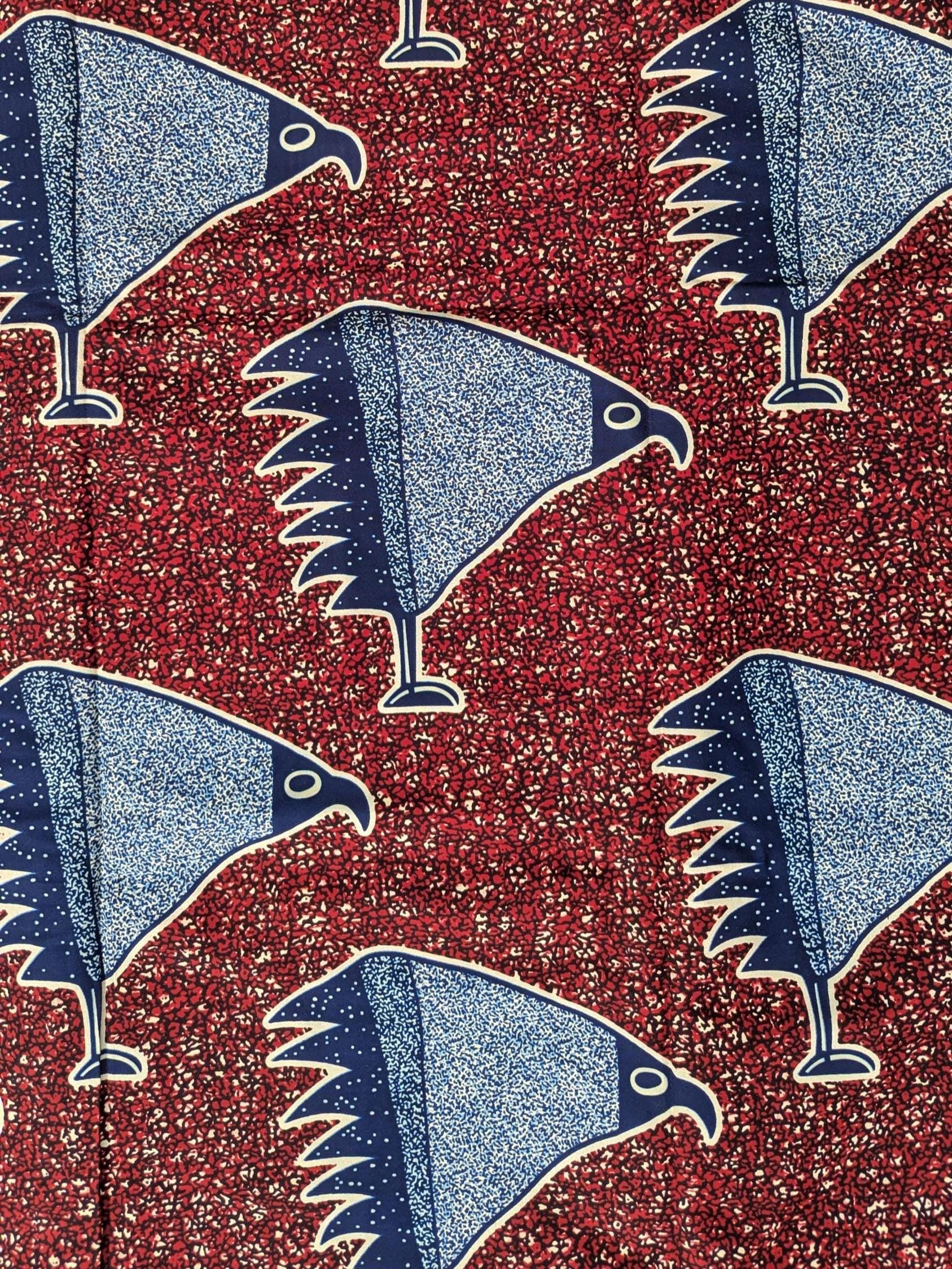African wax print fabric with a striking pattern of stylised blue birds resembling eagles or hawks, each standing on one leg. The birds are illustrated in a speckled texture with navy and sky blue wings, set against a rich red background filled with small, abstract cream and black shapes. Bold and graphic, this fabric is ideal for statement fashion or unique accessories.