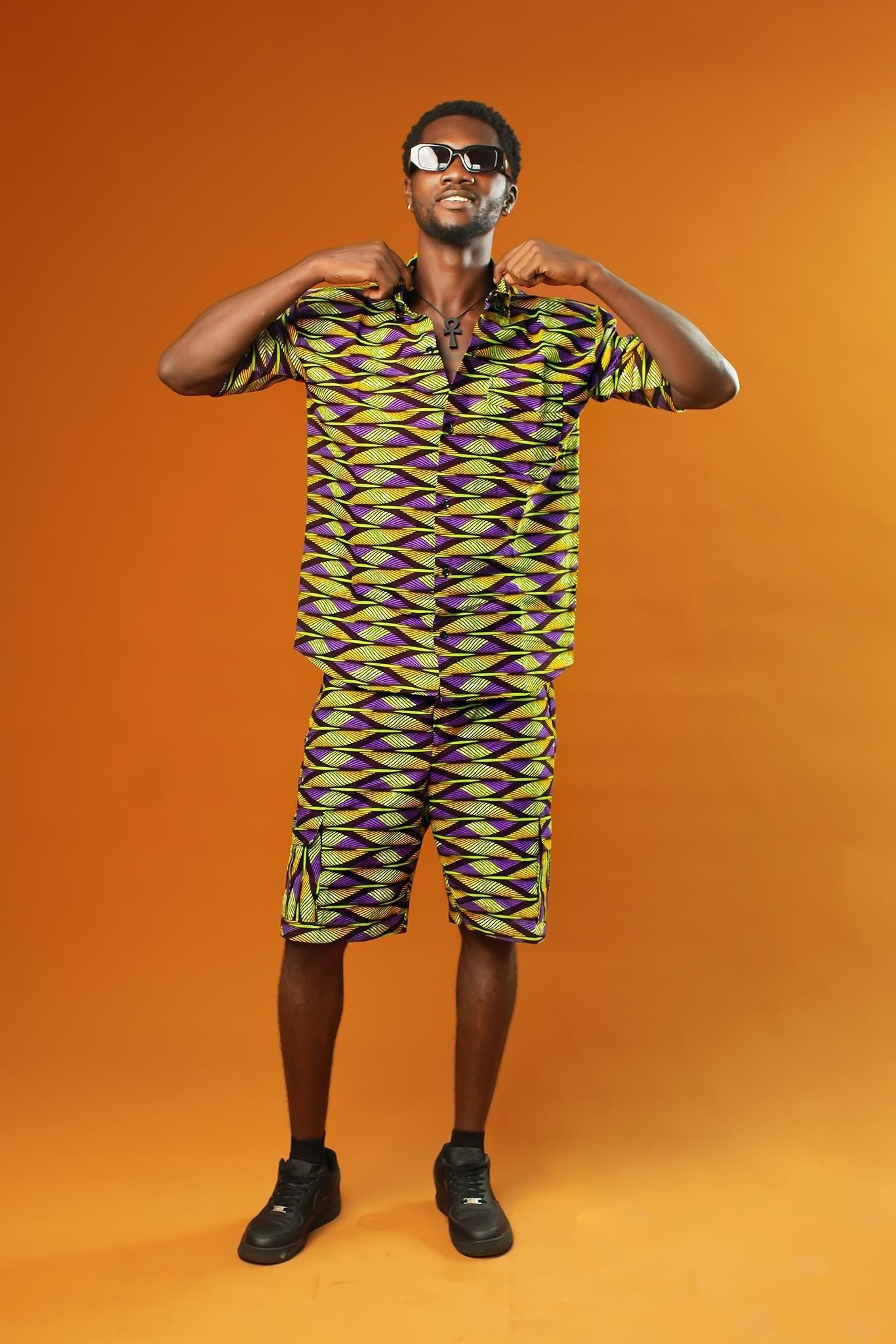Man wearing Aztec green men’s shorts made from African print cotton fabric with a yellow, black, and purple geometric pattern. The shorts are knee-length with a relaxed fit and side cargo pockets. Paired with a matching shirt, black trainers, and sunglasses. Orange background.