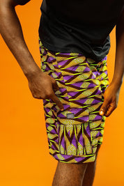Man wearing Aztec green men’s shorts made from African print cotton fabric with a yellow, black, and purple geometric pattern. The shorts are knee-length with a relaxed fit and side cargo pockets. Paired with a matching shirt, black trainers, and sunglasses. Orange background.