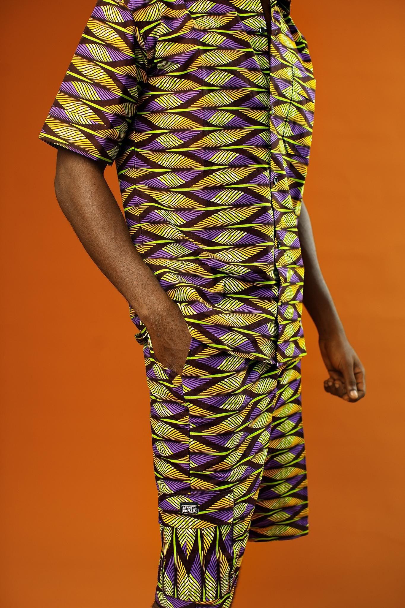 Man wearing Aztec green men’s shorts made from African print cotton fabric with a yellow, black, and purple geometric pattern. The shorts are knee-length with a relaxed fit and side cargo pockets. Paired with a matching shirt, black trainers, and sunglasses. Orange background.