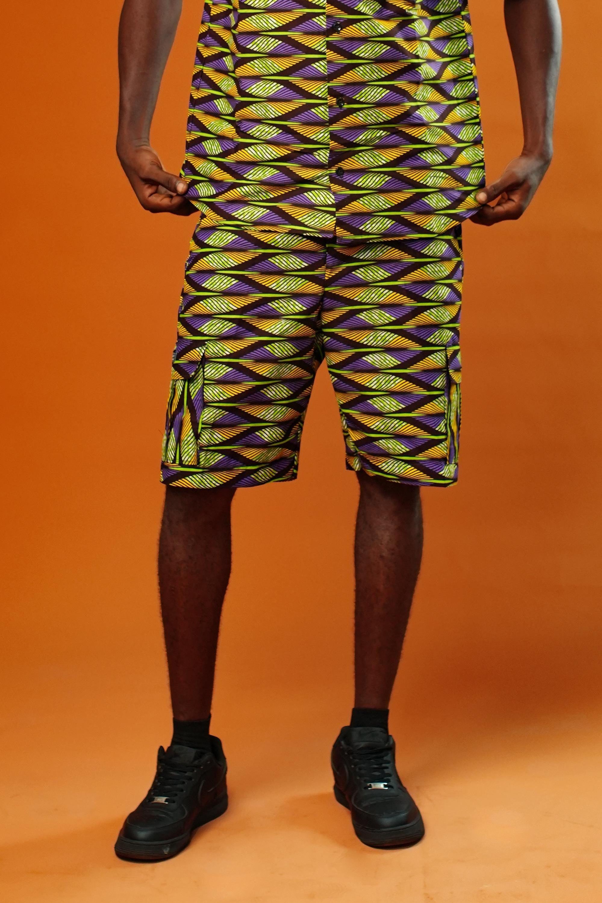 Man wearing Aztec green men’s shorts made from African print cotton fabric with a yellow, black, and purple geometric pattern. The shorts are knee-length with a relaxed fit and side cargo pockets. Paired with a matching shirt, black trainers, and sunglasses. Orange background.