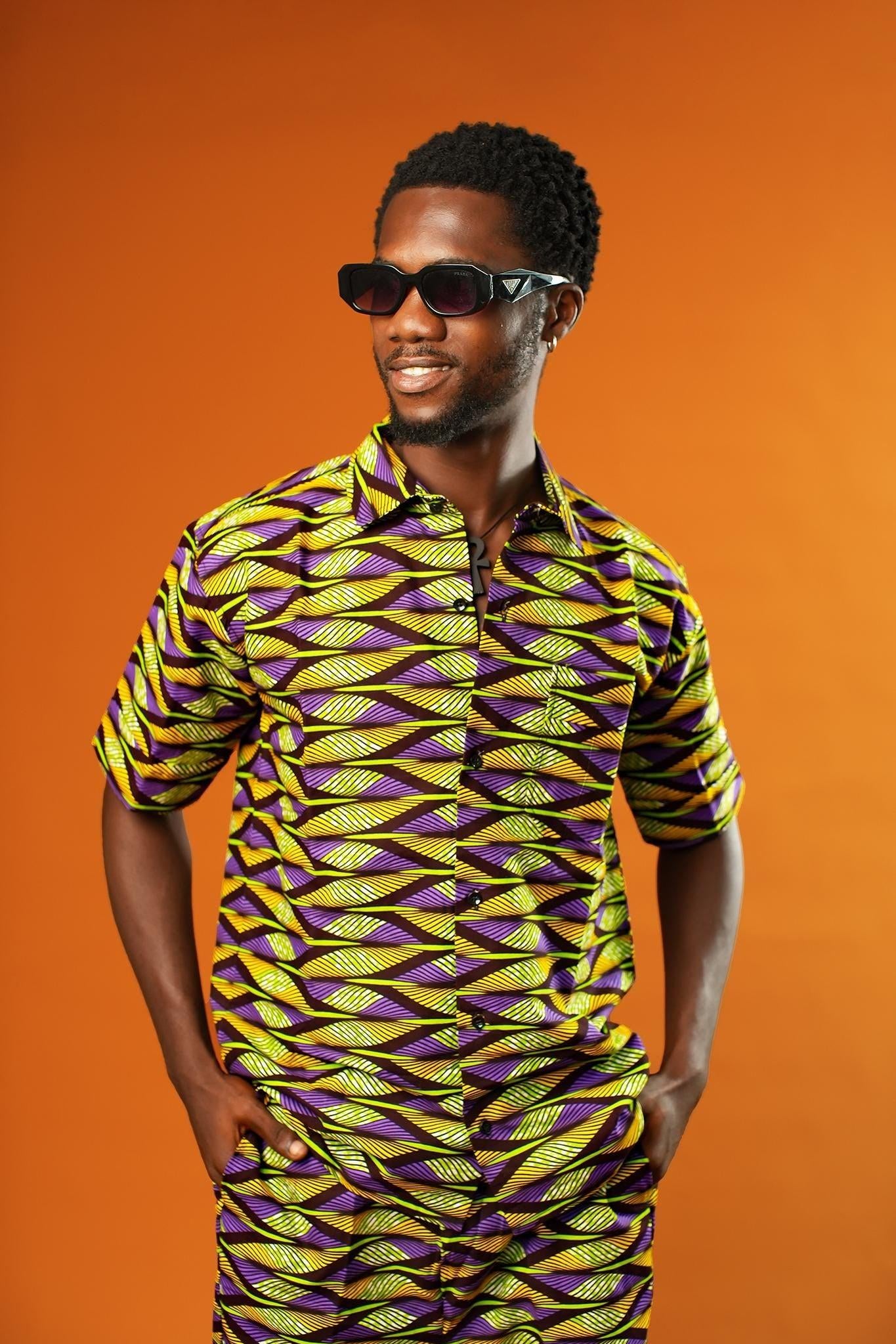 A man strikes a confident pose against a bold orange background, wearing a matching two-piece outfit made from vibrant African wax print fabric. The short-sleeved button-up shirt and shorts feature a striking geometric pattern in neon green, purple, and black.