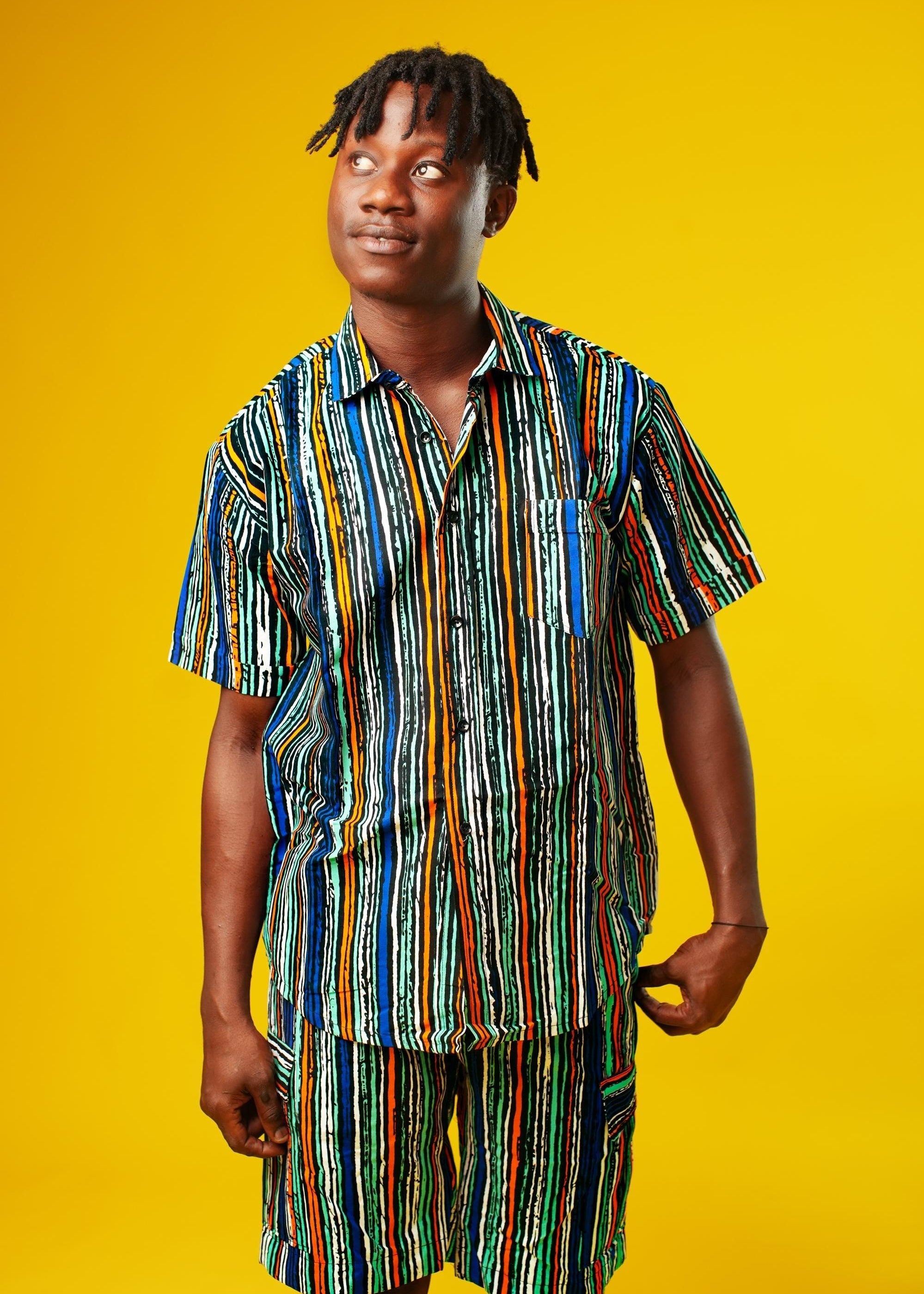 A man walks confidently against a vibrant yellow background wearing a stylish matching two-piece outfit. The set features a short-sleeved button-up shirt and shorts made from a multicoloured striped African print fabric with vertical lines in blue, green, orange, black, and white. He pairs the look with black slide sandals, creating a relaxed yet bold summer-ready vibe.
