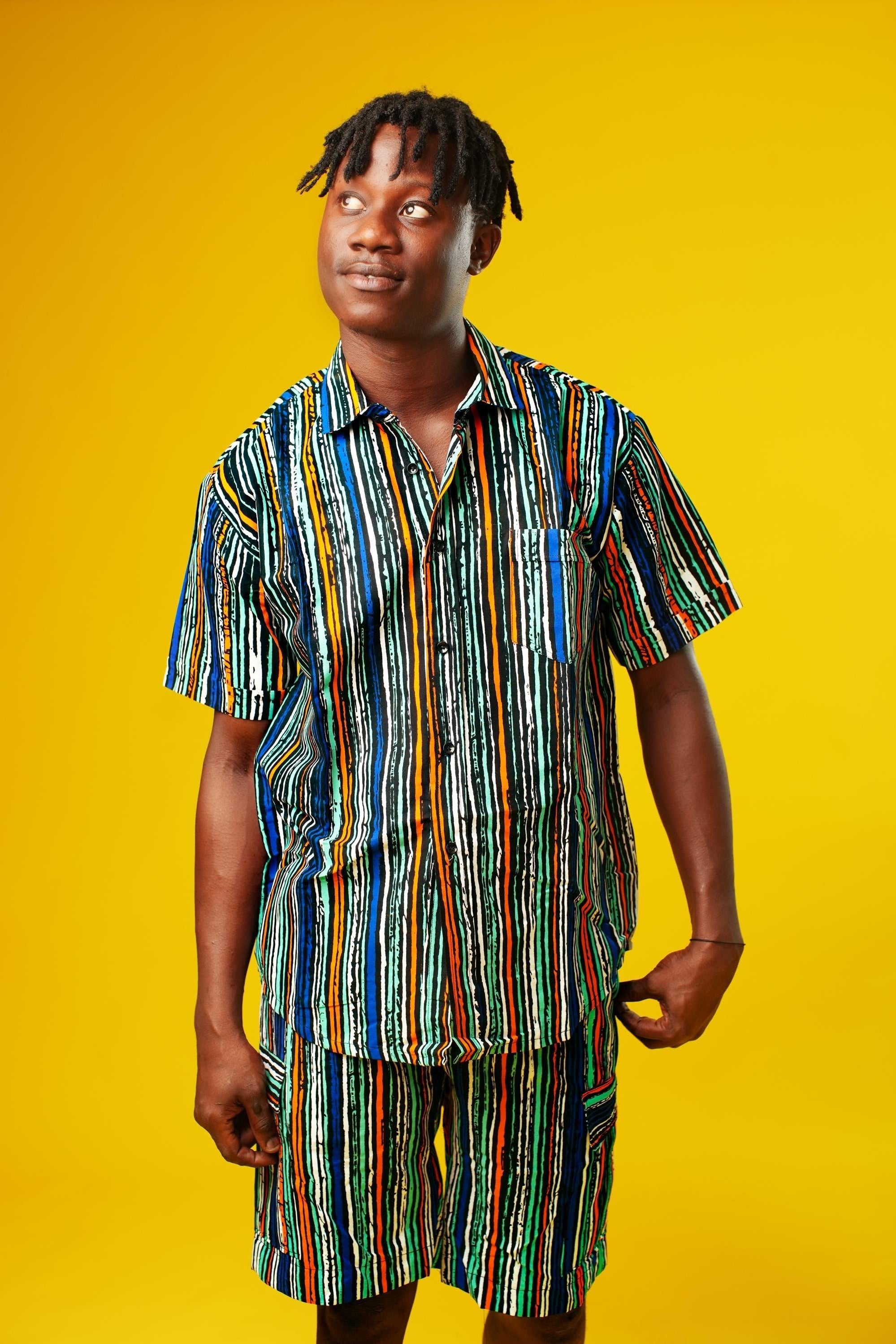 A man walks confidently against a vibrant yellow background wearing a stylish matching two-piece outfit. The set features a short-sleeved button-up shirt and shorts made from a multicoloured striped African print fabric with vertical lines in blue, green, orange, black, and white. He pairs the look with black slide sandals, creating a relaxed yet bold summer-ready vibe.