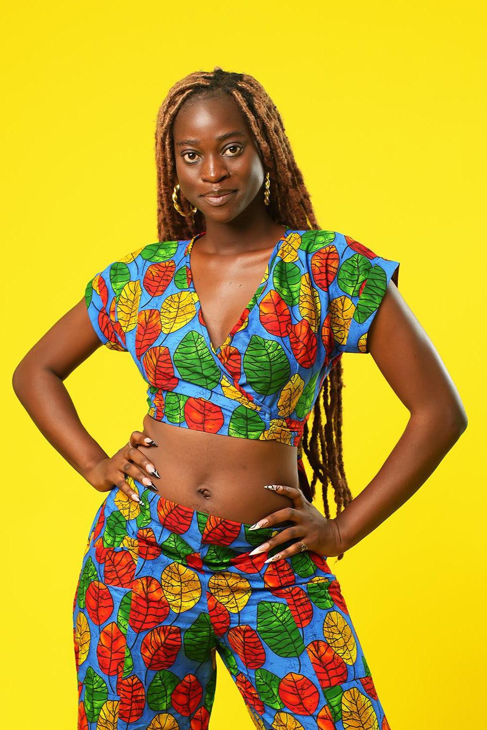 Woman wearing a colourful African print wrap top with a V-neck and short sleeves, made from bright blue fabric featuring red, yellow, and green leaf patterns. The top has a cropped fit and ties at the waist.