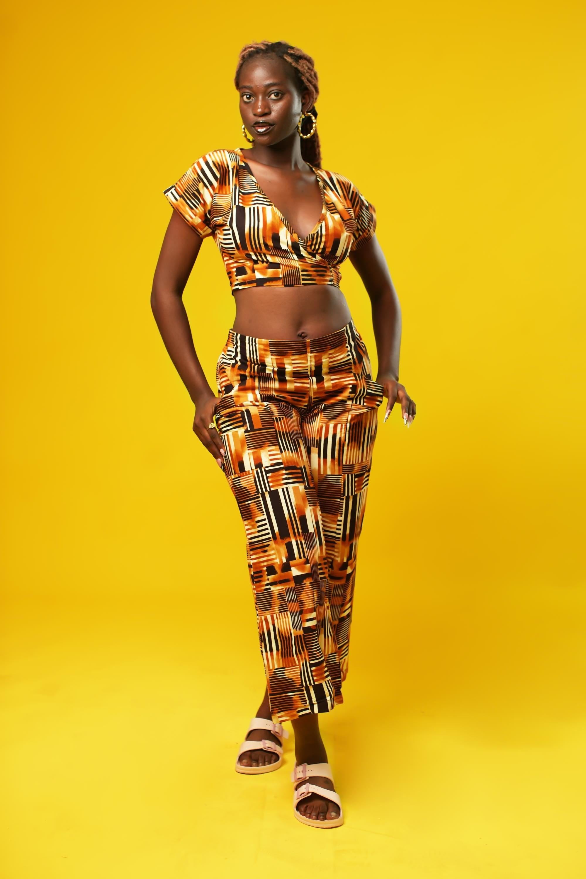 Woman wearing wide leg African palazzo pants with a geometric brown, yellow, orange, and black print. The cropped trousers are made from lightweight cotton, styled as part of a festival outfit with a matching wrap crop top.