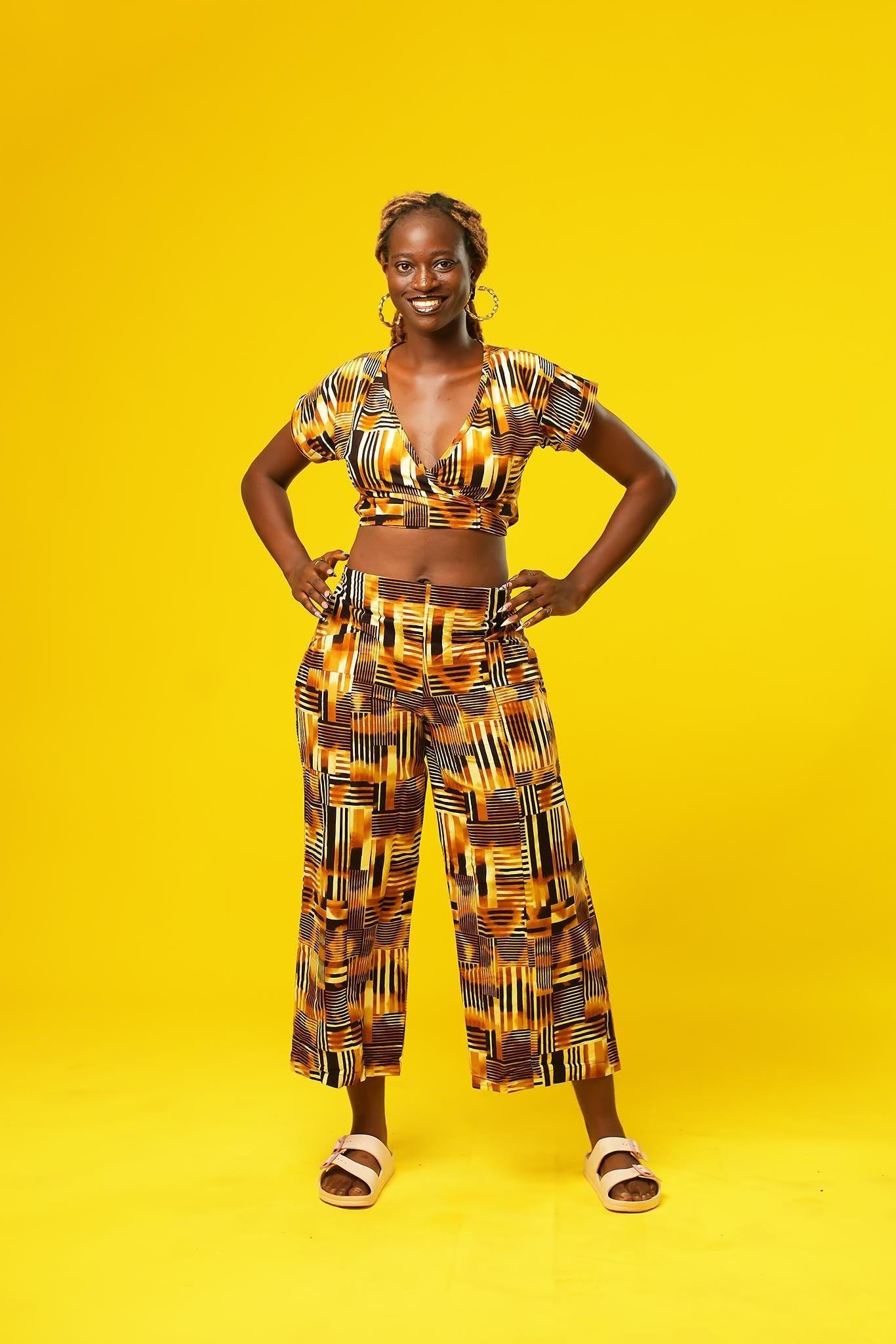 Woman wearing wide leg African palazzo pants with a geometric brown, yellow, orange, and black print. The cropped trousers are made from lightweight cotton, styled as part of a festival outfit with a matching wrap crop top.