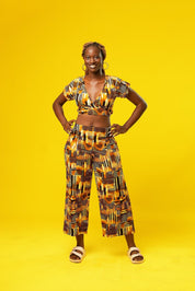 Woman wearing wide leg African palazzo pants with a geometric brown, yellow, orange, and black print. The cropped trousers are made from lightweight cotton, styled as part of a festival outfit with a matching wrap crop top.