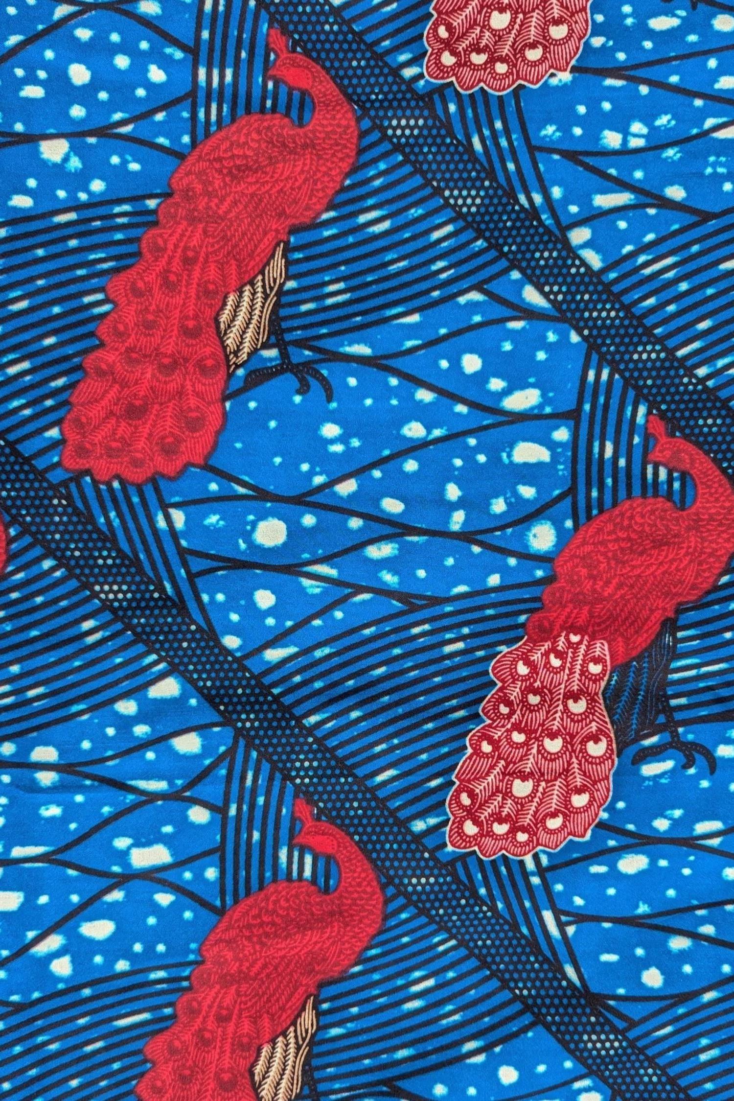 Bright and bold African wax print fabric featuring elegant pink peacocks with intricately patterned tail feathers standing against a vivid blue background. The design includes diagonal black lines and abstract wave-like patterns with splashes of cream, creating a striking contrast and sense of movement. Perfect for standout fashion pieces or statement accessories.
