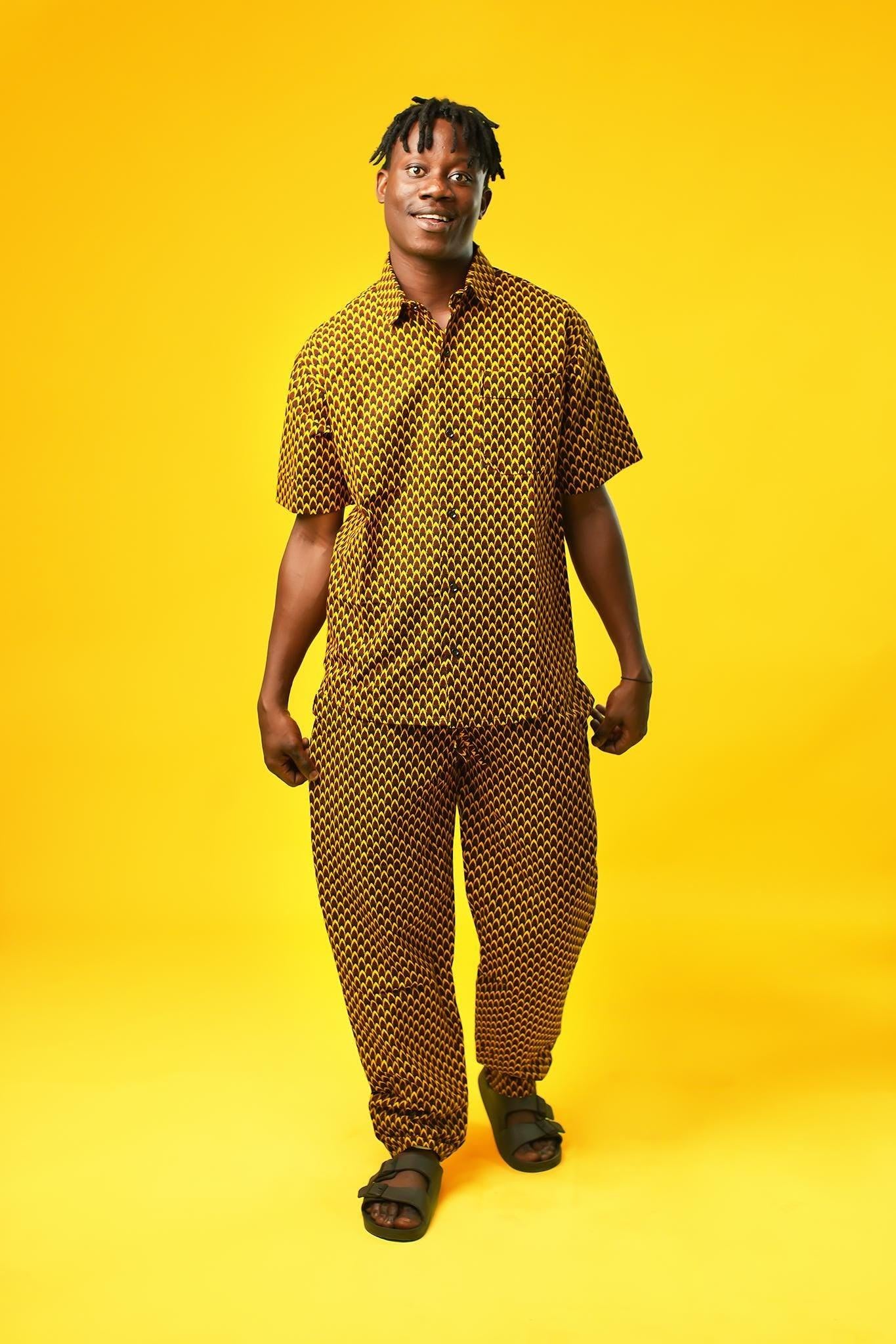 A man poses against a vibrant yellow background wearing a coordinated two-piece outfit made from a rich mustard yellow and black African print fabric. The set includes a short-sleeved button-up shirt and relaxed drawstring trousers featuring a bold geometric pattern.
