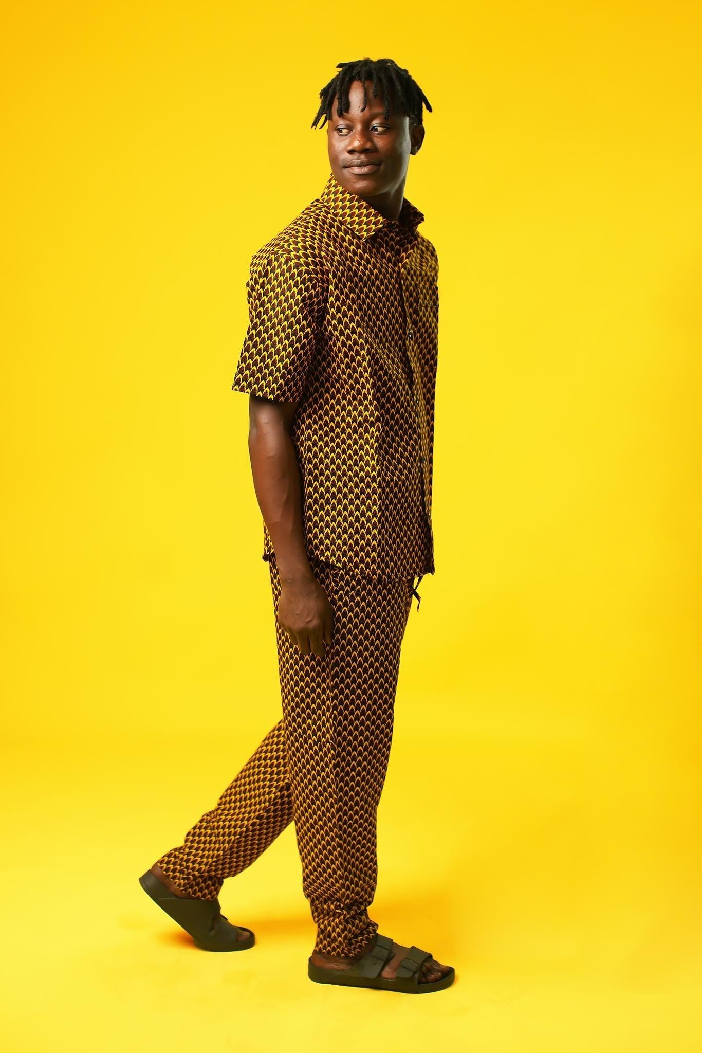 A man poses against a vibrant yellow background wearing a coordinated two-piece outfit made from a rich mustard yellow and black African print fabric. The set includes a short-sleeved button-up shirt and relaxed drawstring trousers featuring a bold geometric pattern.