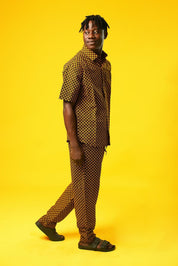 A man poses against a vibrant yellow background wearing a coordinated two-piece outfit made from a rich mustard yellow and black African print fabric. The set includes a short-sleeved button-up shirt and relaxed drawstring trousers featuring a bold geometric pattern.