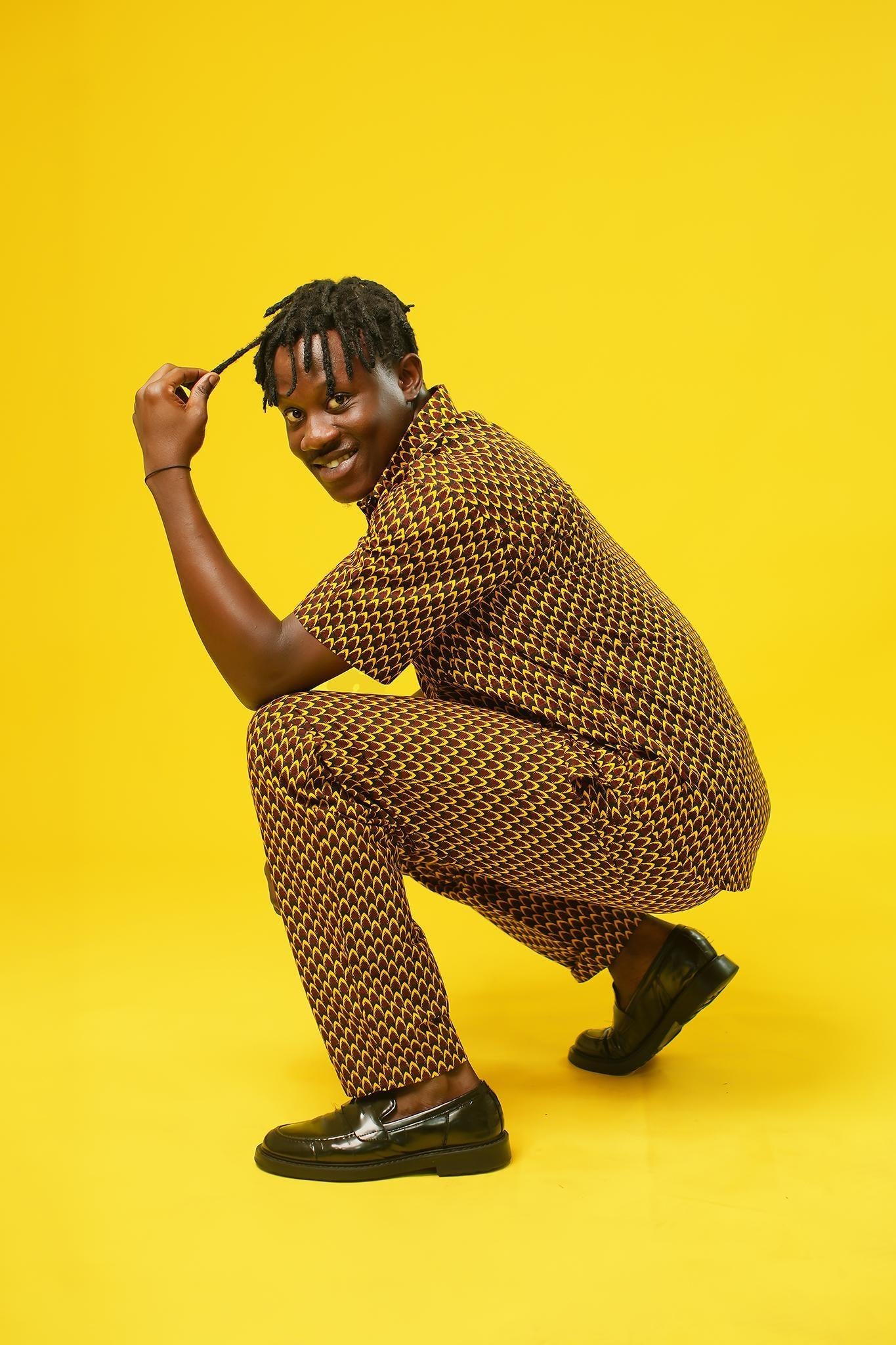 A man poses against a vibrant yellow background wearing a coordinated two-piece outfit made from a rich mustard yellow and black African print fabric. The set includes a short-sleeved button-up shirt and relaxed drawstring trousers featuring a bold geometric pattern.