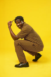 A man poses against a vibrant yellow background wearing a coordinated two-piece outfit made from a rich mustard yellow and black African print fabric. The set includes a short-sleeved button-up shirt and relaxed drawstring trousers featuring a bold geometric pattern.