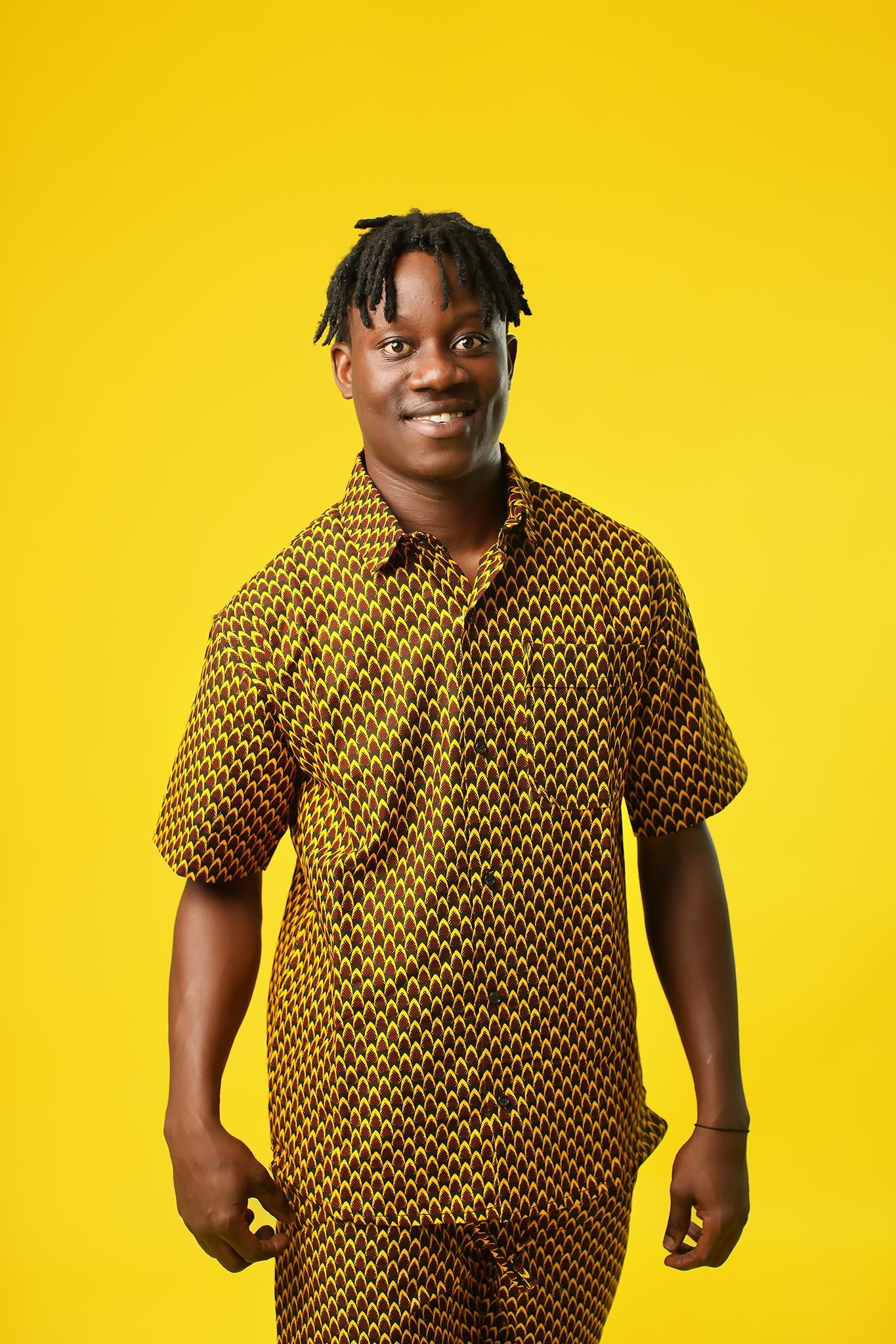 A man poses against a vibrant yellow background wearing a coordinated two-piece outfit made from a rich mustard yellow and black African print fabric. The set includes a short-sleeved button-up shirt and relaxed drawstring trousers featuring a bold geometric pattern.