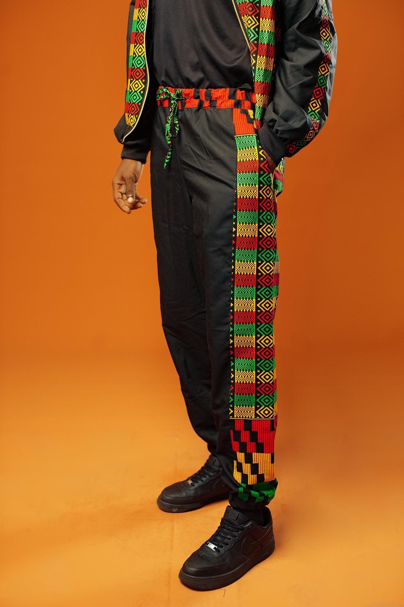 Man standing against an orange background wearing black tracksuit trousers with colourful Kente African print panels down the sides. The trousers have a relaxed fit with elastic cuffs at the ankles and a drawstring waist.
