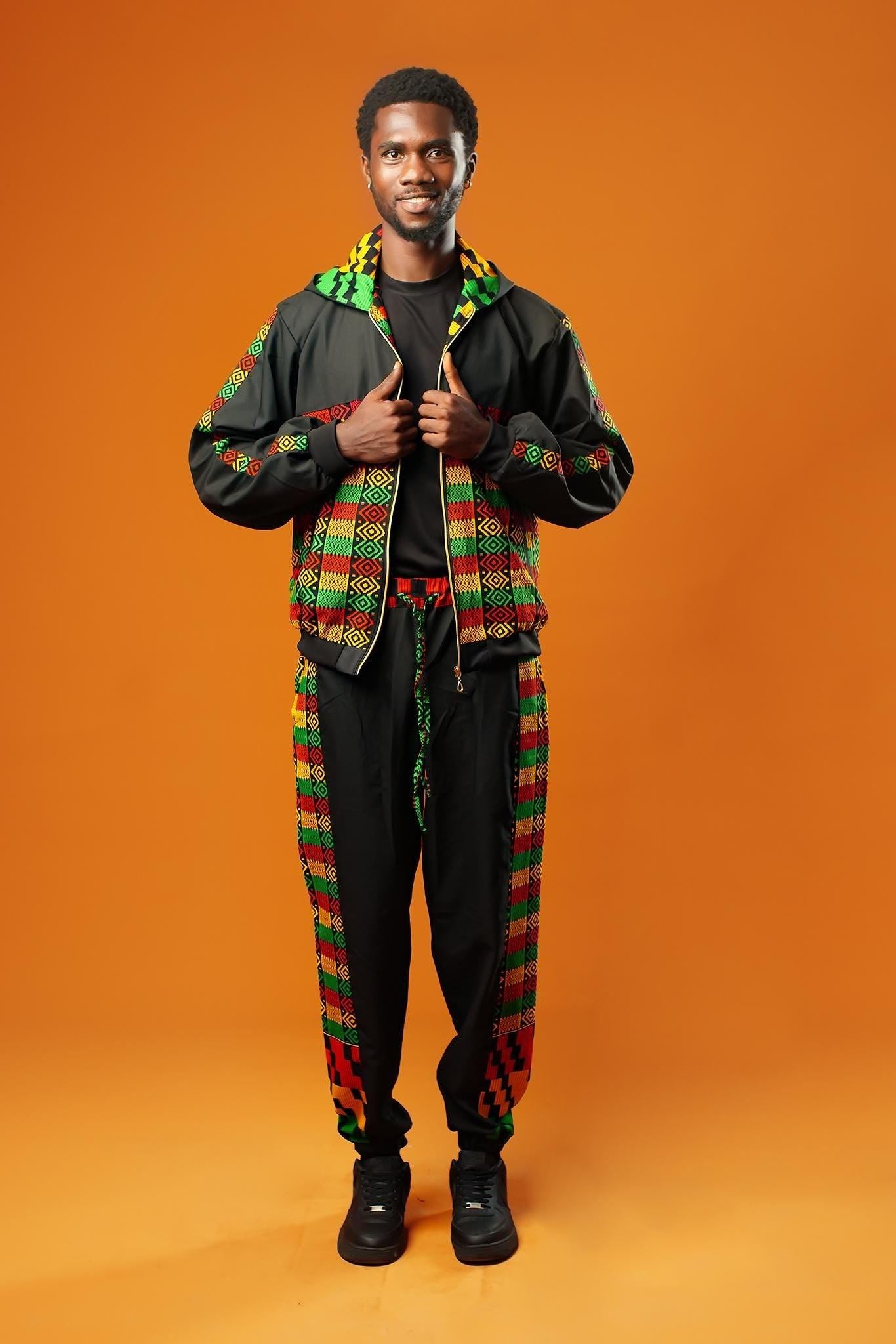 Man standing against an orange background wearing black tracksuit trousers with colourful Kente African print panels down the sides. The trousers have a relaxed fit with elastic cuffs at the ankles and a drawstring waist.