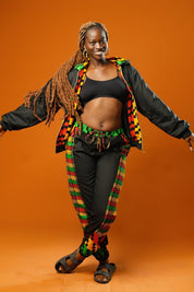 Woman standing against an orange background wearing black tracksuit trousers with colourful Kente African print panels down the sides. The trousers have a relaxed fit with elastic cuffs at the ankles and a drawstring waist. She pairs them with a matching black jacket featuring Kente trim and a black crop top.