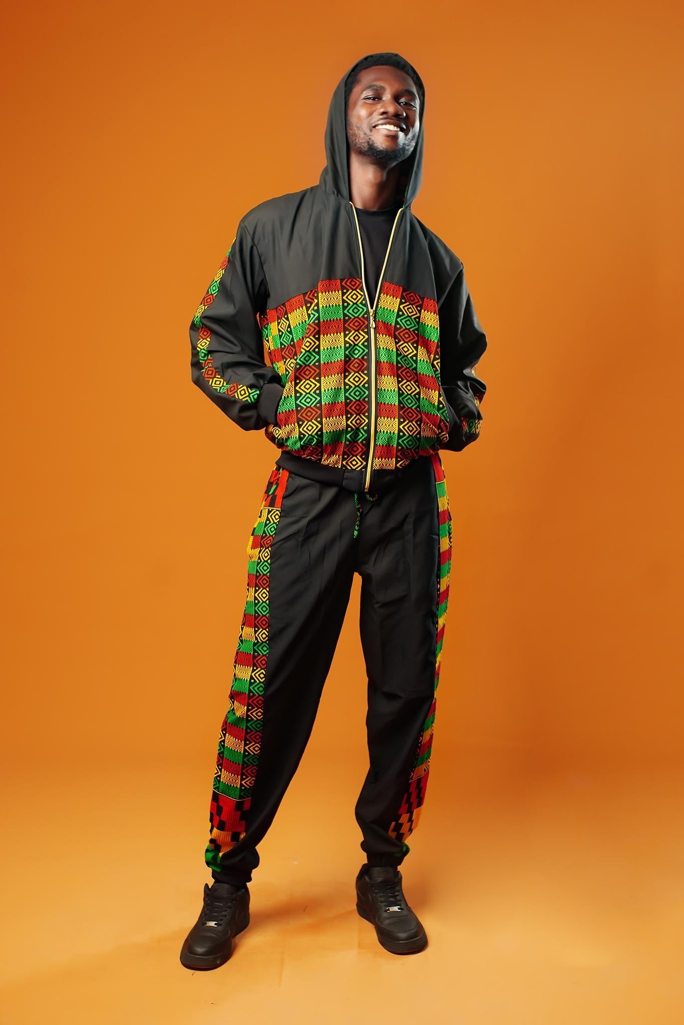 Man standing against an orange background wearing black tracksuit trousers with colourful Kente African print panels down the sides. The trousers have a relaxed fit with elastic cuffs at the ankles and a drawstring waist.