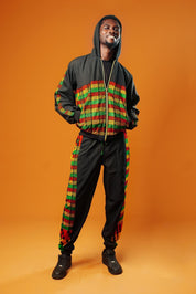 Man standing against an orange background wearing black tracksuit trousers with colourful Kente African print panels down the sides. The trousers have a relaxed fit with elastic cuffs at the ankles and a drawstring waist.
