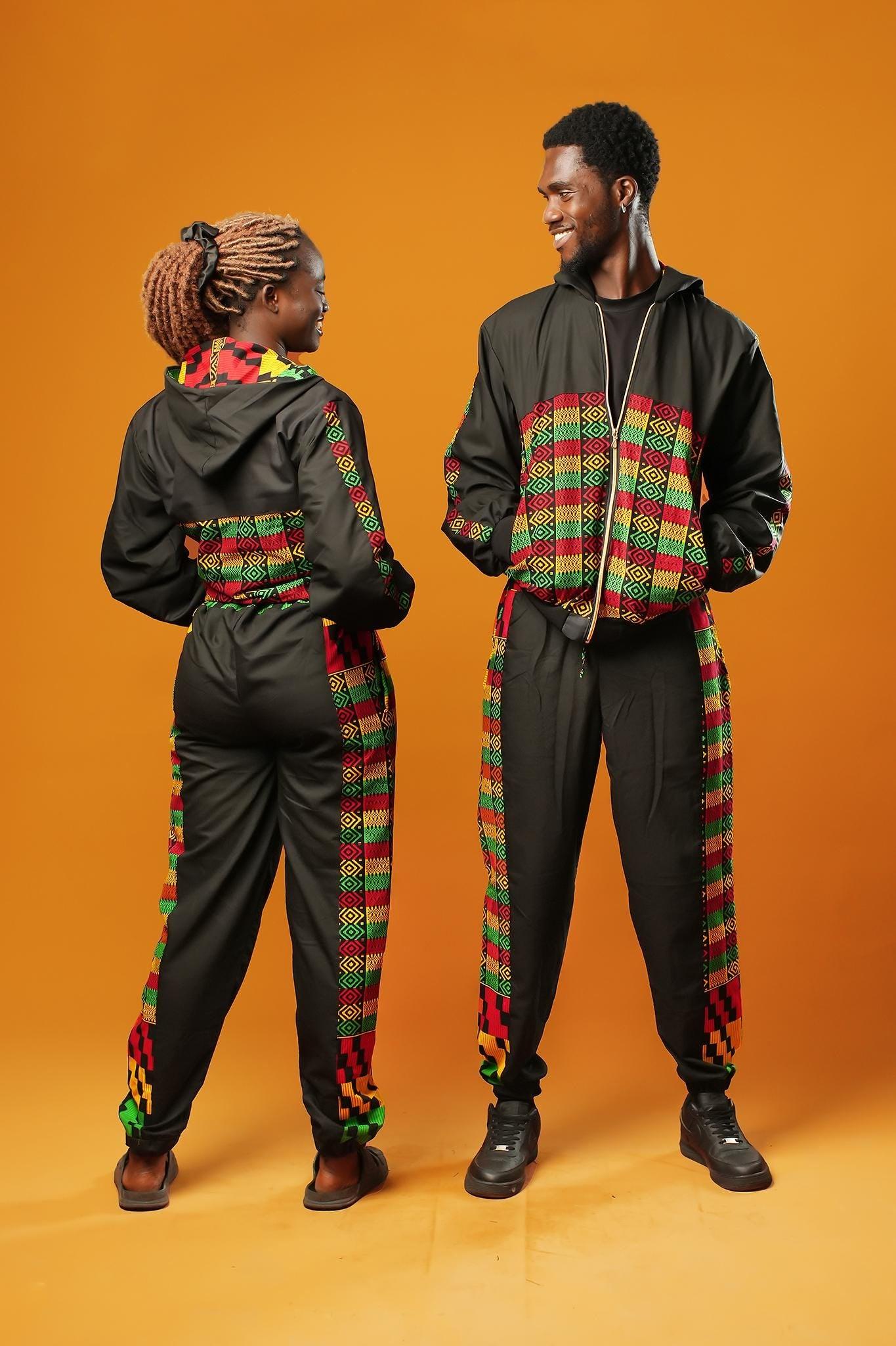 Woman and man standing against an orange background wearing black tracksuit trousers with colourful Kente African print panels down the sides. The trousers have a relaxed fit with elastic cuffs at the ankles and a drawstring waist. They both pair them with matching black jackets featuring Kente trim.
